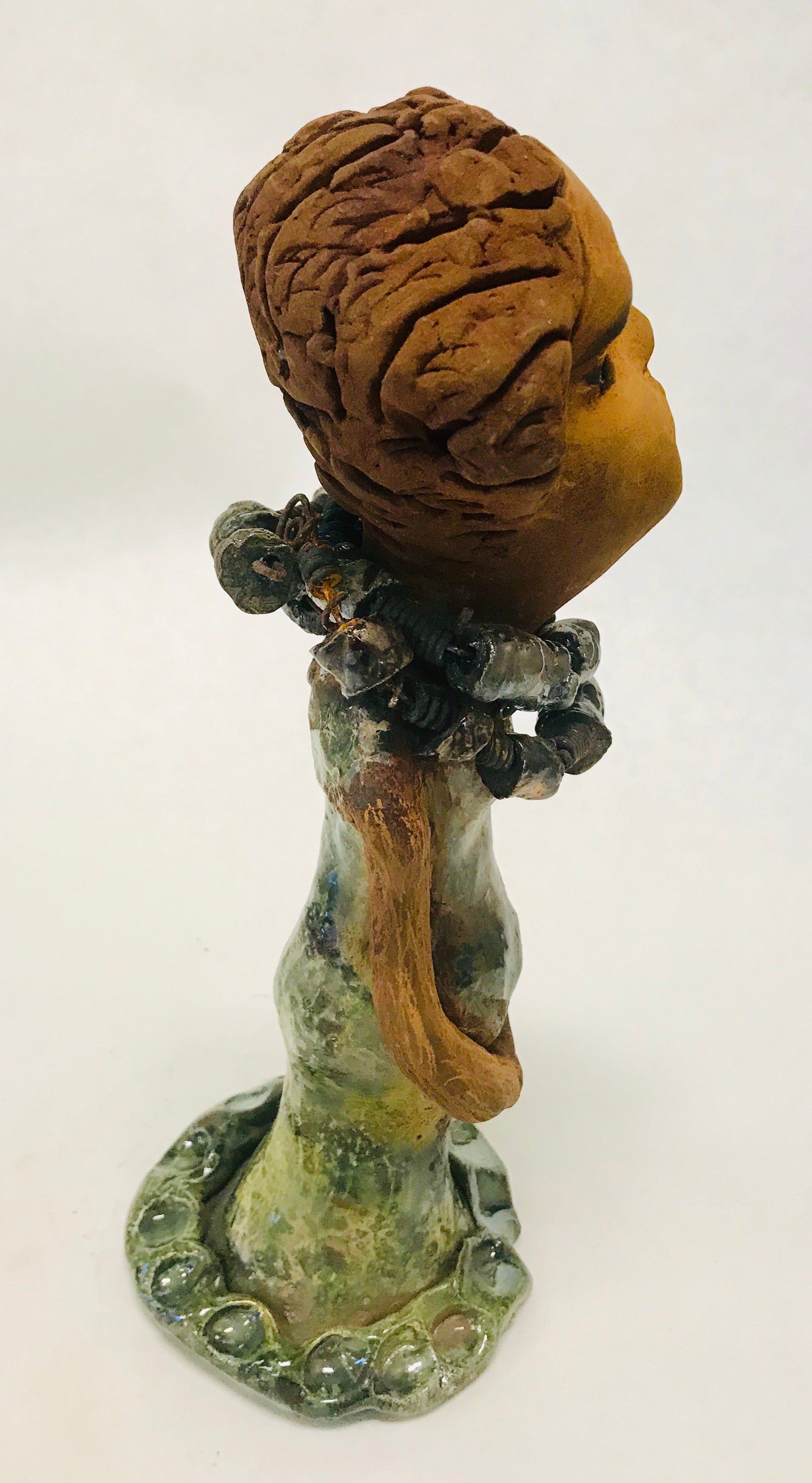Greta stands 11" x 4" x 4" and weighs 2.5 lbs. She has a lovely honey brown complexion with  reddish brown lips. She has a braided hairstyle.  Greta has a colorful metallic green antique copper glazed dress. She wears two raku beaded necklaces. With long eyes slightly opened, Greta has hopes of finding a new home.    Greta will attract and spark conversation with guest in your home!