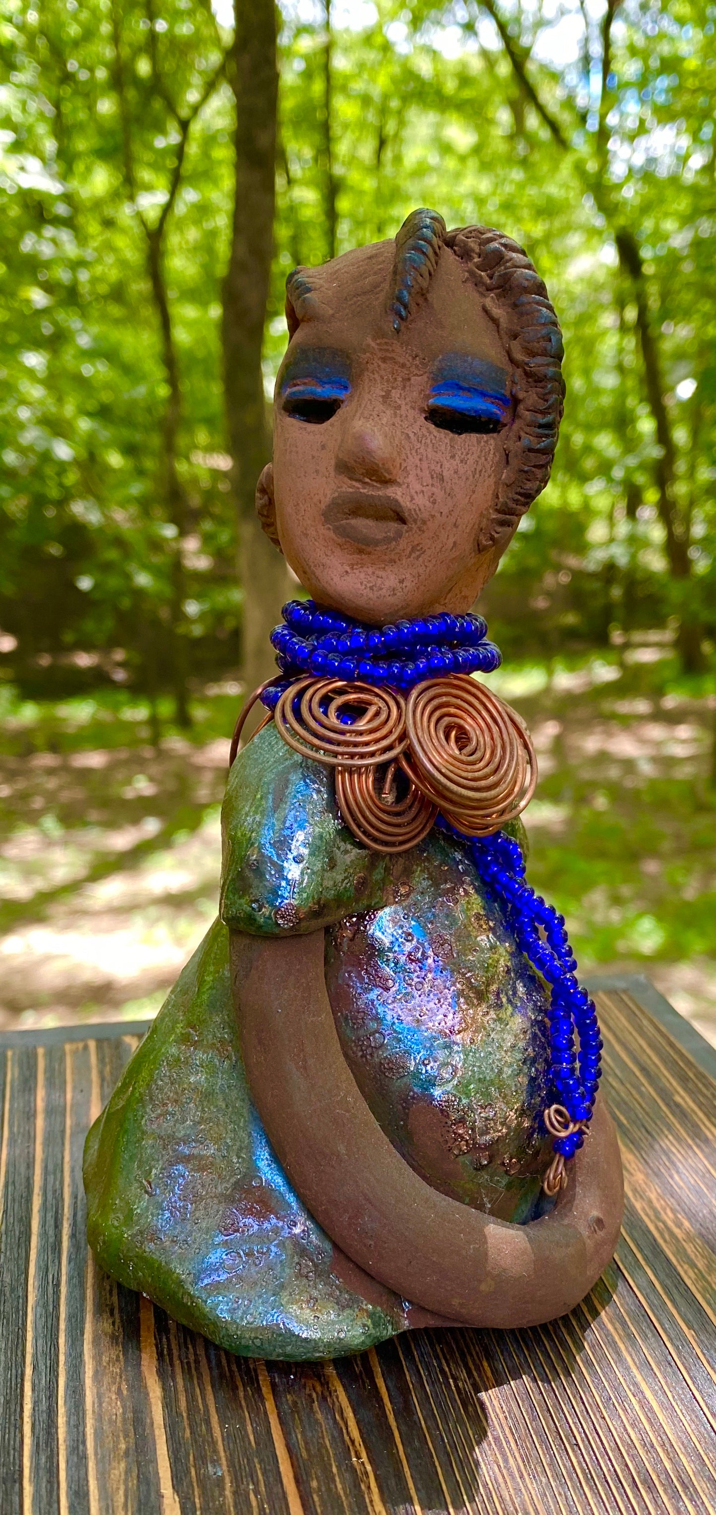 Meet Ife! Ife stands 7.5" x 4" x 5" and weighs 1.02 lbs. She has a lovely honey brown complexion with reddish brown lips. She has a short braided hairstyle. Ife has a colorful metallic antique copper glazed dress. She wears spiral copper wire necklaces on top of an aqua blue beaded collar. Ife long loving arms rest at her side. With accent blue eye shadow and eyes wide opened, Ife has hopes of finding a new home.