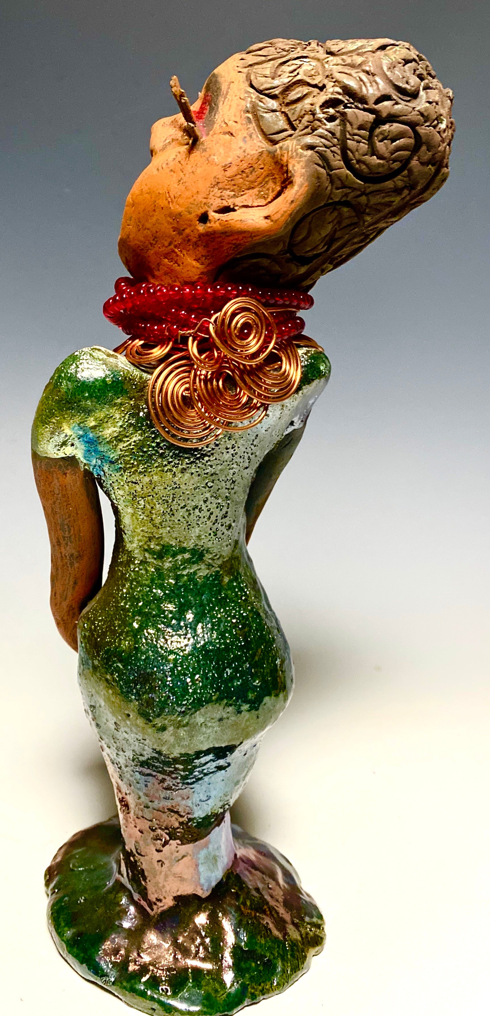 Meet Mona! Mona stands 11.5" x 4" x 4" and weighs 1.81 lbs. She has a lovely chocolate brown complexion with chocolate brown lips. She has a braided hairstyle.  Mona has a colorful metallic antique copper glazed dress. She wears a spiral copper wire necklaces on top of a beaded ruby red collar. With long lashes and eyes wide opened, Mona has hopes of finding a new home. You may have noticed, Mona is the one of a few herdew  sculptures that wears a face mask during this unprecedented time.