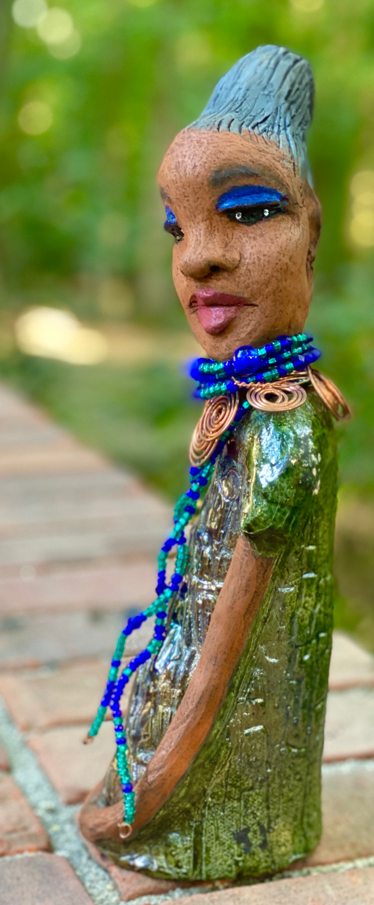 Meet Emma Again! Emma went and got her a makeover too. She was jealous of Eonna! Emma stands 12.5" x 3. 5" x 4" and weighs 2.04 lbs. She has a lovely honey brown complexion with reddish brown lips. She has a braided clay hairstyle. Emma has a colorful metallic green antique copper glazed dress. She wears a spiral copper wire necklace on top of a multicolored blue beaded collar. Check out Emma before and after pics. With eyes wide opened, Emma has hopes of finding her way into your home!