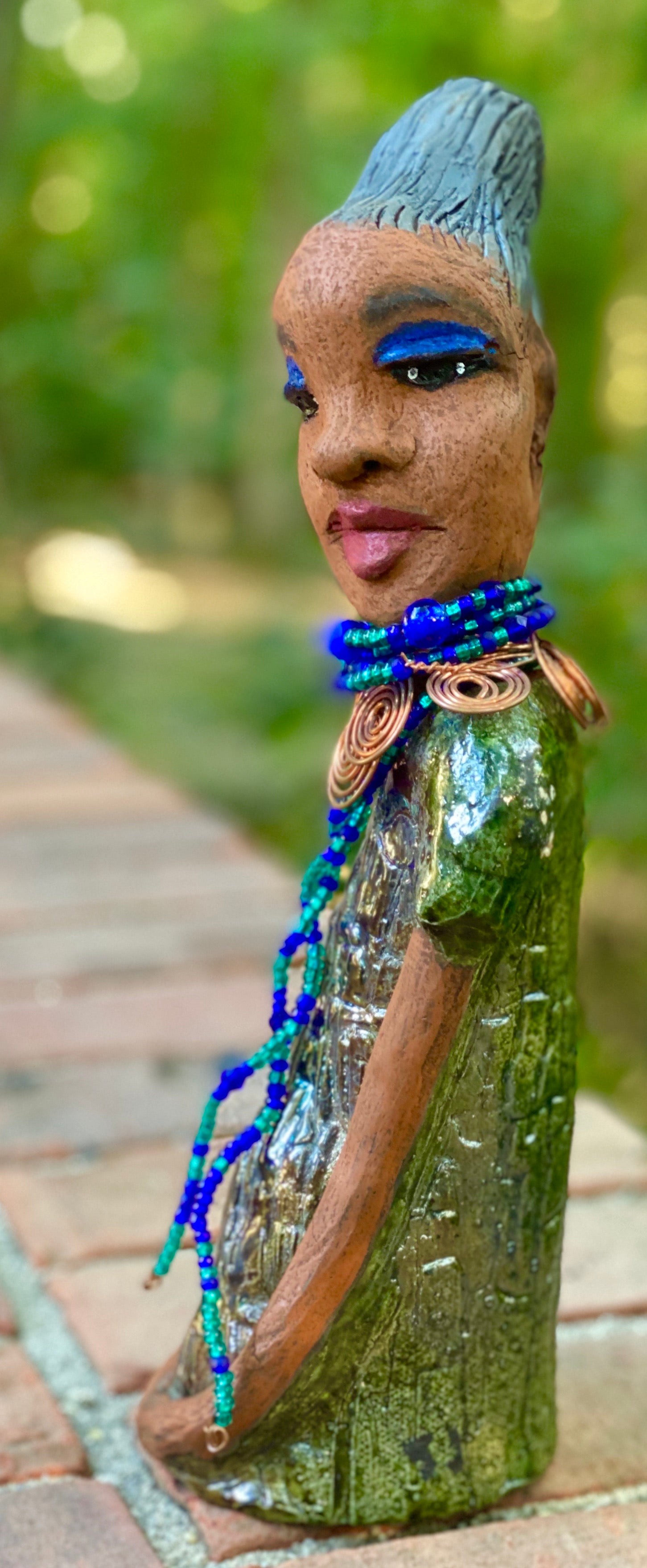 Meet  Emma Again! Emma went and got her a makeover too. She was jealous of Eonna! Emma stands 12.5" x 3. 5" x 4" and weighs 2.04 lbs. She has a lovely honey brown complexion with reddish brown lips. She has a braided clay hairstyle.  Emma has a colorful metallic green antique copper glazed dress. She wears a spiral copper wire necklace on top of a multicolored blue beaded collar. Check out Emma before and after pics. With eyes wide opened,  Emma has hopes of finding  her way into your home!