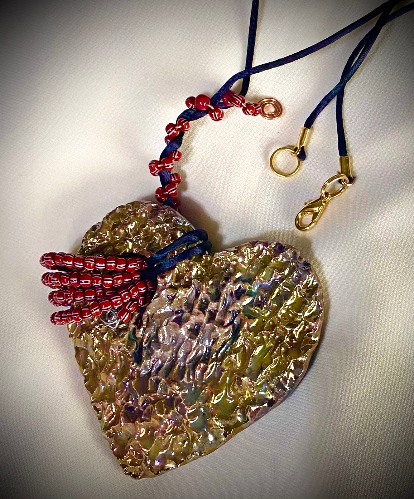 Have A Heart ! Each heart pendant is handmade with love! It is 3"x 3" and weighs approx. 3ozs. This pendant has a white and gold metallic raku glazes that renders a unique translucent patina. The heart has a textured with cut out cross pattern . It holds a spiral of multicolored mini beads on a spiral copper wire. This pendant has a nice 12" black suede cord!