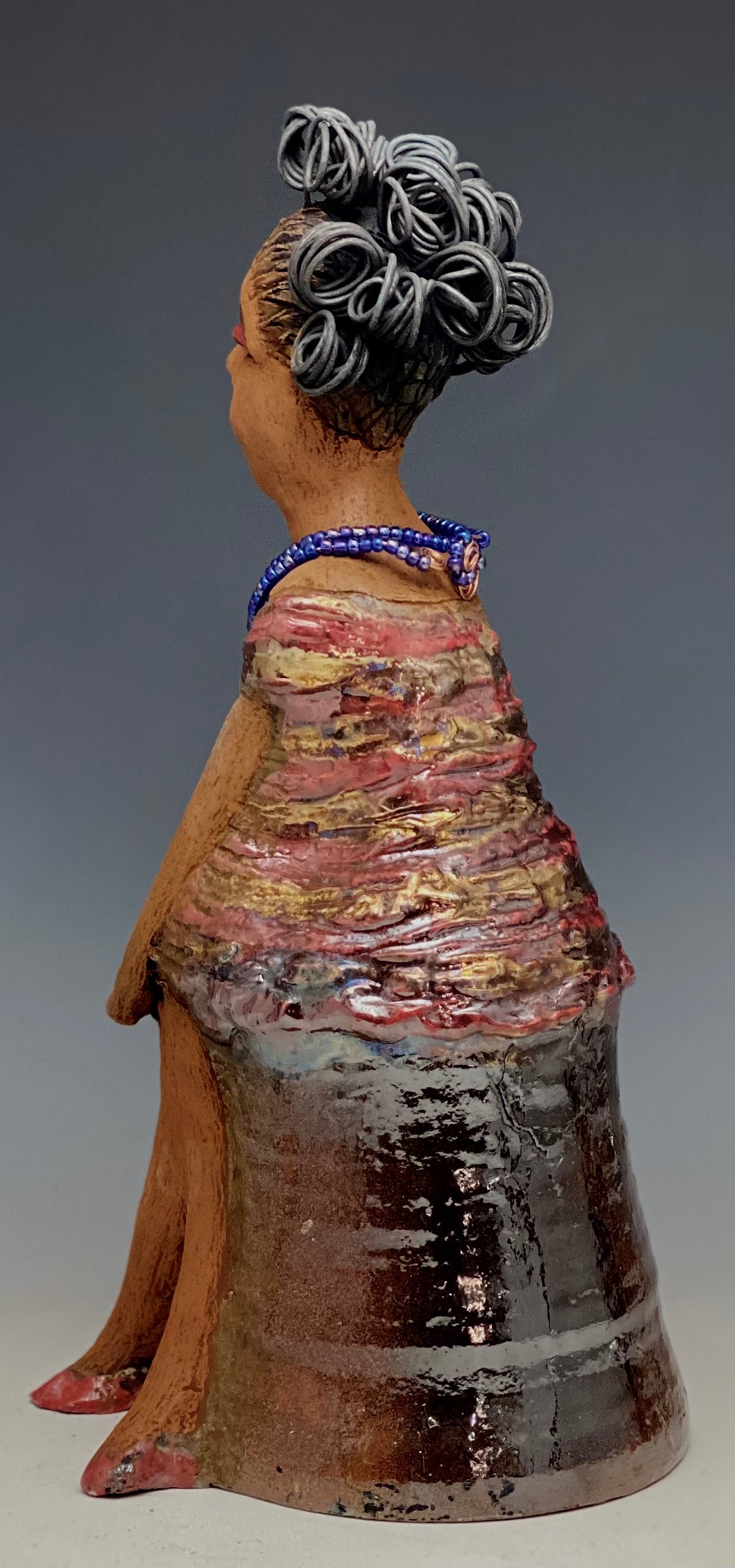 You know someone like Kim. One who reaps elegance and sophistication! Kim stands 13" x 5" x 7" and weighs 2.07 lbs. She has a lovely light beige complexion with over 4 feet of 16 gauge spiral coiled wire hair. Kim's metallic copper red and gold dress is accented by a multicolored aqua blue beaded necklace. Kim's maroon eye shadow gives her a distinguished mystical look. One must wonder what is Kim thinking? Kim sits with her legs crossed as she awaits a special place to grace your home.