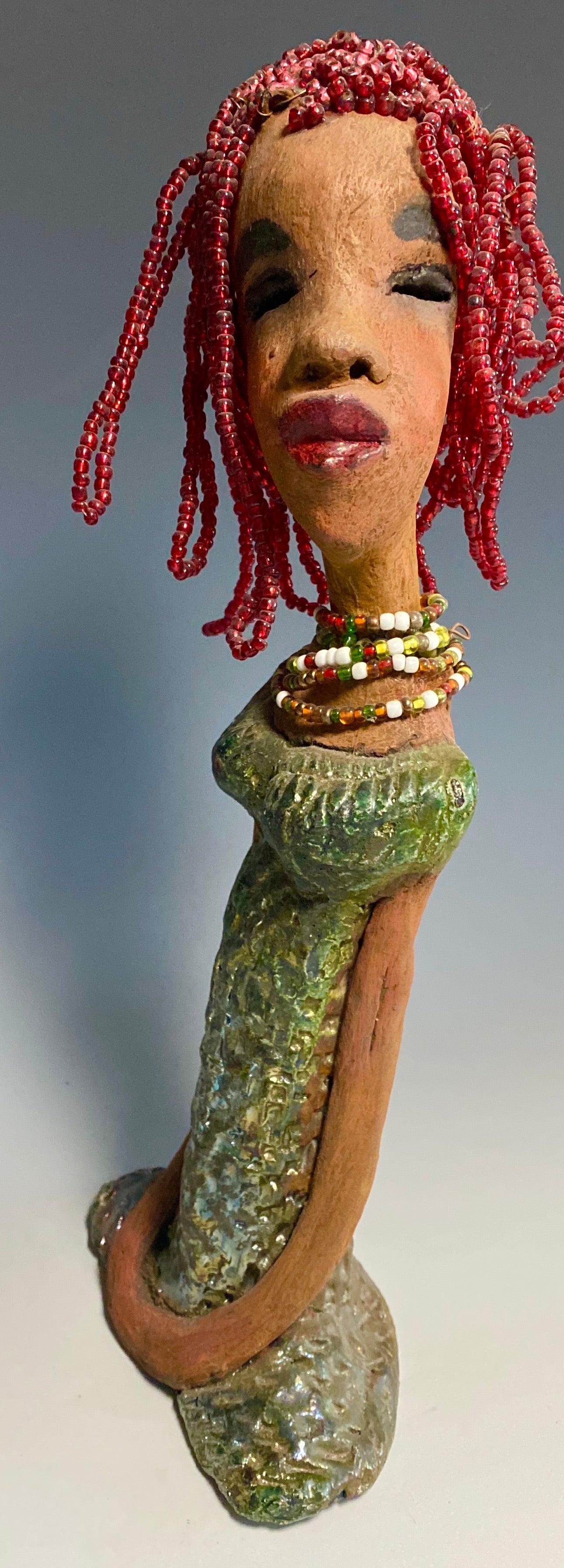 Meet July! July stands 14" x 5" x 3" and weighs 2.05 lbs. July has a head full of red beaded hair. She has a honey brown complexion. July dress has a metallic textured copper green.  She wears a multi colored beaded necklace. With her head slightly turned her long loving arms gently rest at her side.