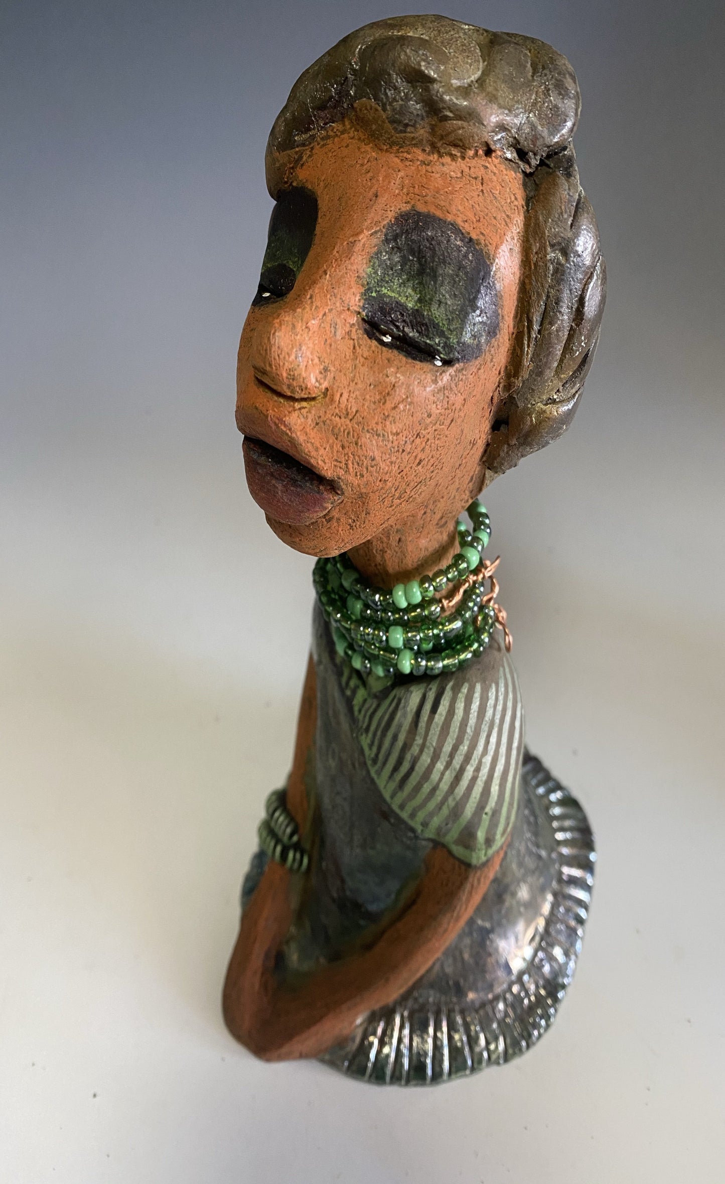 Meet Cora! Cora stands 9" x 5" x 6" and weighs 1.12 lbs. She has a lovely beige brown complexion with long etched braided clay hair. Cora’s long loving arms rest beside her copper green dress. Her dress is alligator green with a green coiled arm band. Cora wears a long string mixed green beaded necklace. Cora is a sophisticated lady that will grace your home.