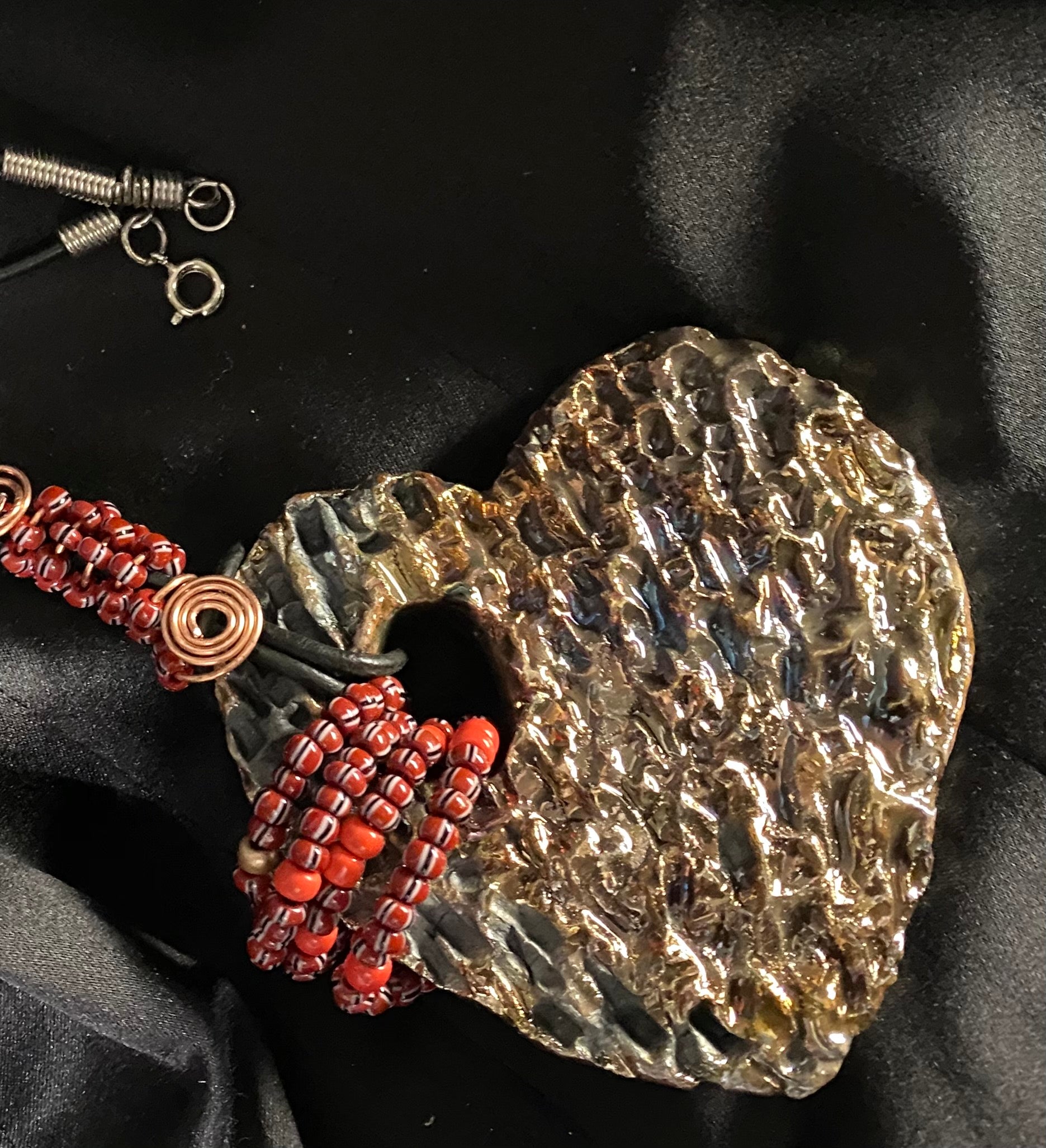 Have A Heart ! Each heart pendant is handmade with love! It is 3"x 3" and weighs approx. 3ozs. This pendant has a gold metallic raku glazes that renders a unique translucent  patina. The heart has a textured  pattern. It holds a spiral of ruby mini beads on a spiral copper wire. This pendant has a nice 12" black suede cord!