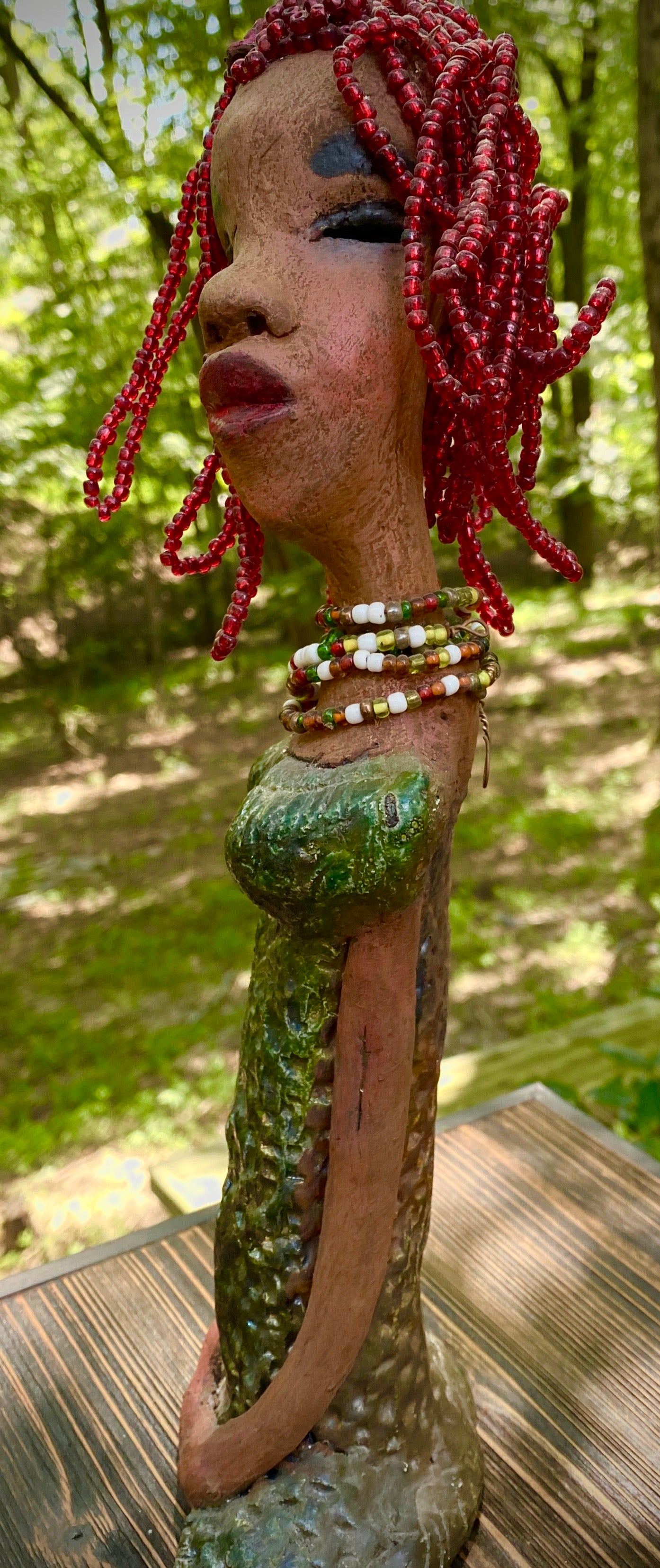 Meet July! July stands 14" x 5" x 3" and weighs 2.05 lbs. July has a head full of red beaded hair. She has a honey brown complexion. July dress has a metallic textured copper green.  She wears a multi colored beaded necklace. With her head slightly turned her long loving arms gently rest at her side.