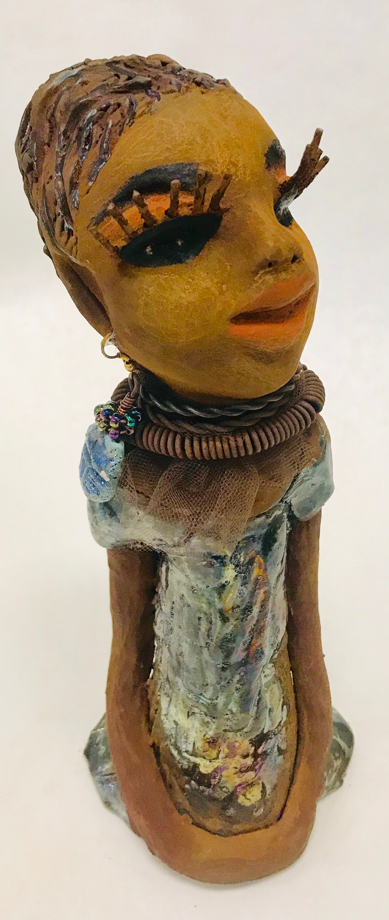 Sorbari stands 11" x 4" x 4" and weighs 2.5 lbs. She has a lovely honey brown complexion with  reddish brown lips. She has a braided hairstyle.  Sorbari has a colorful metallic green antique copper glazed dress. She wears two spiral wire necklaces. With long eyes slightly opened, Sorbari has hopes of finding a new home.