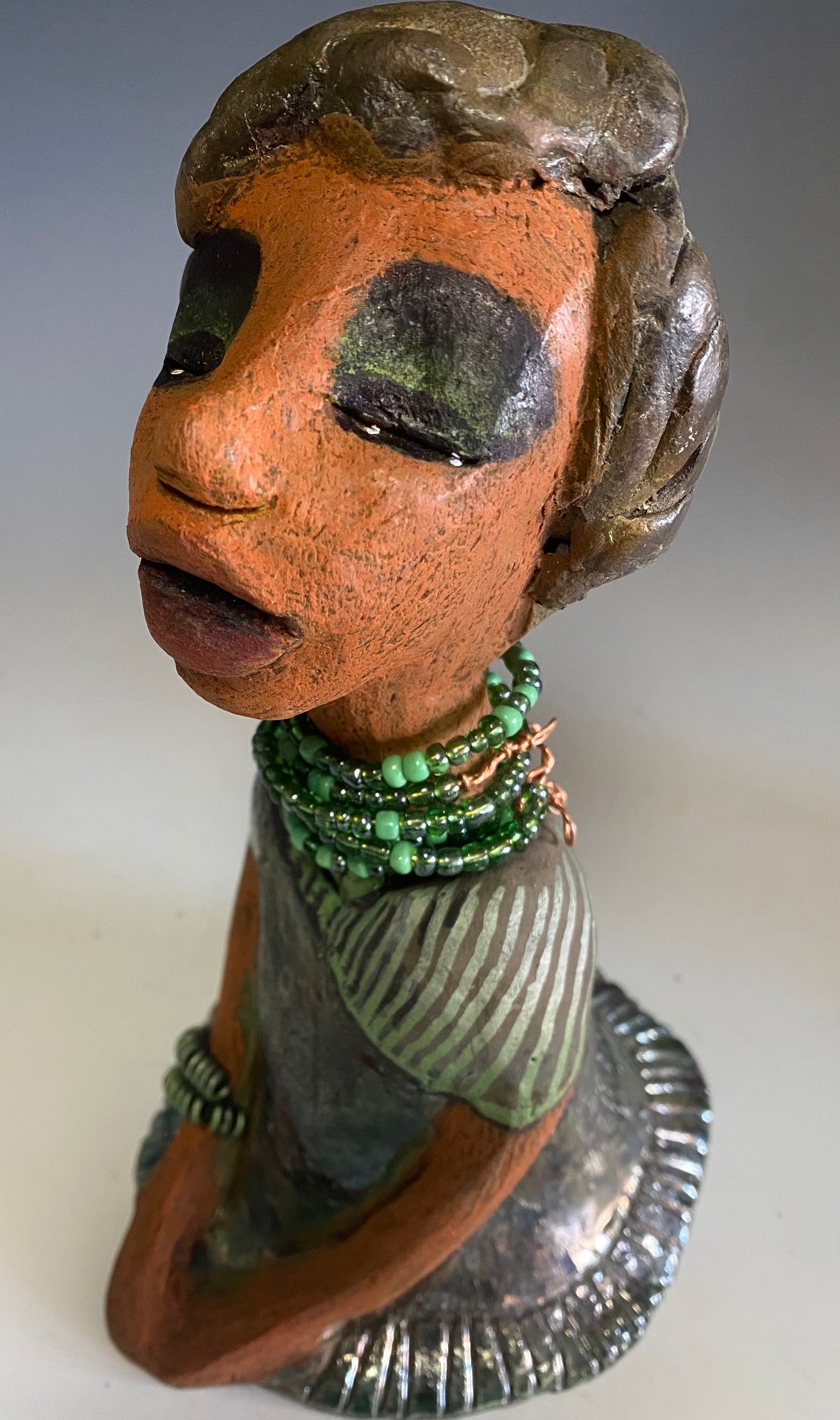 Meet Cora! Cora stands 9" x 5" x 6" and weighs 1.12 lbs. She has a lovely beige brown complexion with long etched braided clay hair. Cora’s long loving arms rest beside her copper green dress. Her dress is alligator green with a green coiled arm band. Cora wears a long string mixed green beaded necklace. Cora is a sophisticated lady that will grace your home.