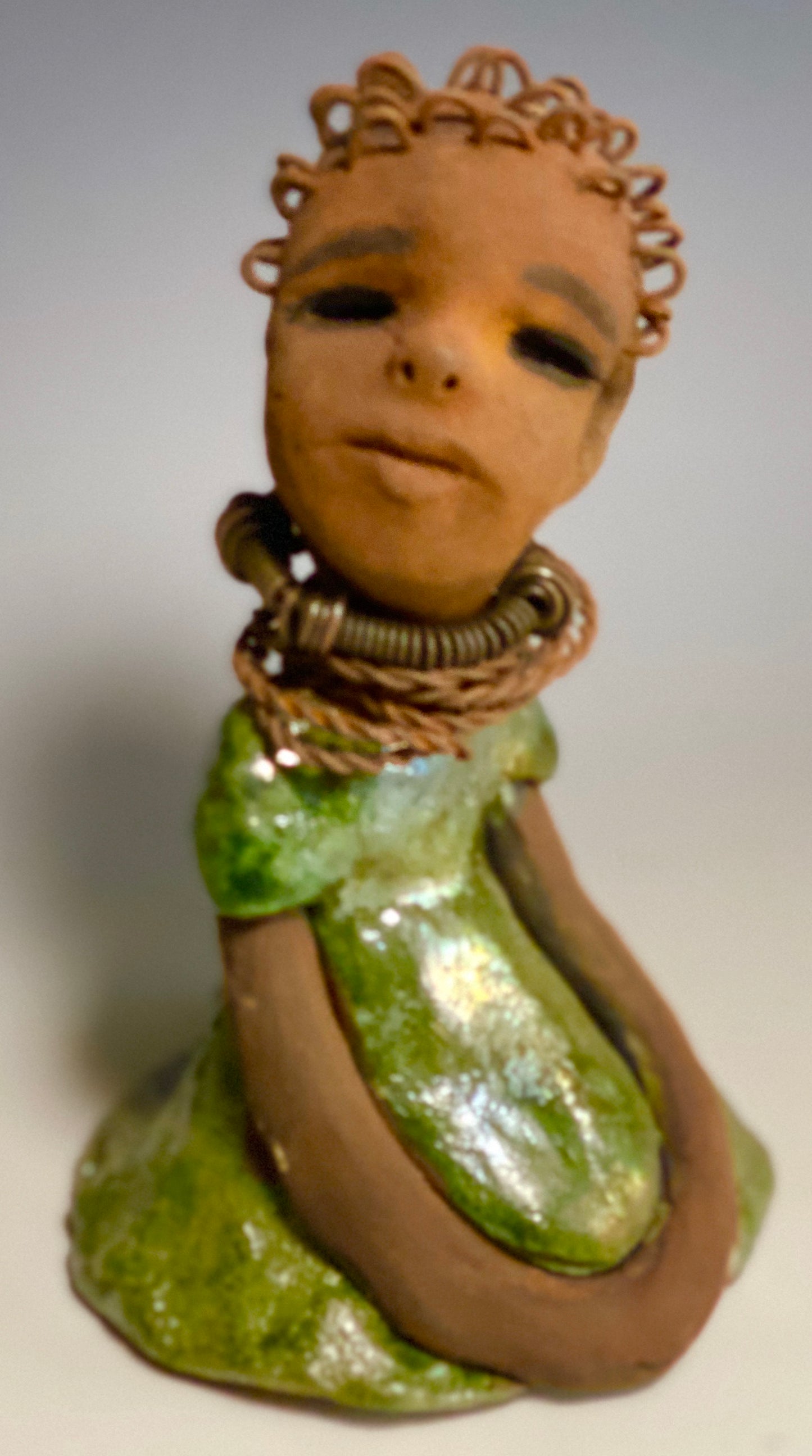 Meet Nura! Nura stands 7.5" x 5" x 5" and weighs 1.06 lbs. She has a lovely honey brown complexion with reddish brown lips. She has a wire braided hairstyle. Nura has a colorful metallic green antique copper glazed dress. She wears spiral wire necklaces With her eyes wide opened, Nura has hopes of finding her way into your home.