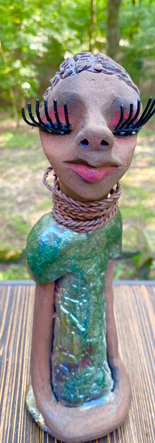 Meet  Ingrid Ingrid stands 12" x 5" x 4" and weighs 1.14 lbs. She has a lovely honey brown complexion with  reddish brown lips. She has a braided hairstyle.  Ingrid has a colorful metallic green antique copper glazed dress. She wears six spiral wire necklaces.  Her long loving arms rest at her sides. With long lashes and eyes slightly opened, Ingrid has hopes of finding a new home!