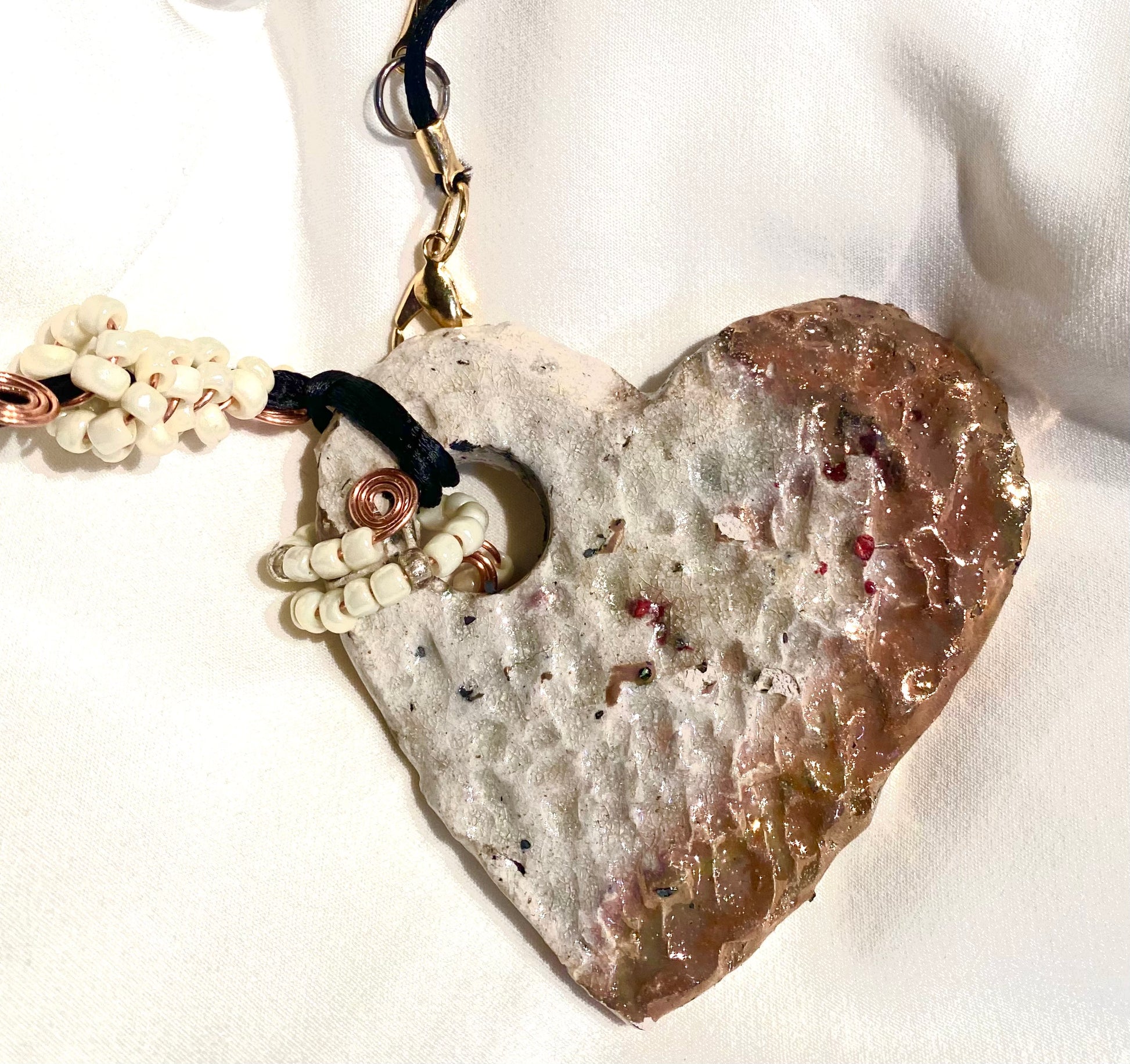 Have A Heart ! Each heart pendant is handmade with love! It is 3" x 3" and weighs approx. 3ozs. It has white metallic raku glazes that renders a unique translucent  patina. The heart is textured and holds a  spiral of white pony beads on copper wire.  This pendant has a nice 12"black rattail cord!