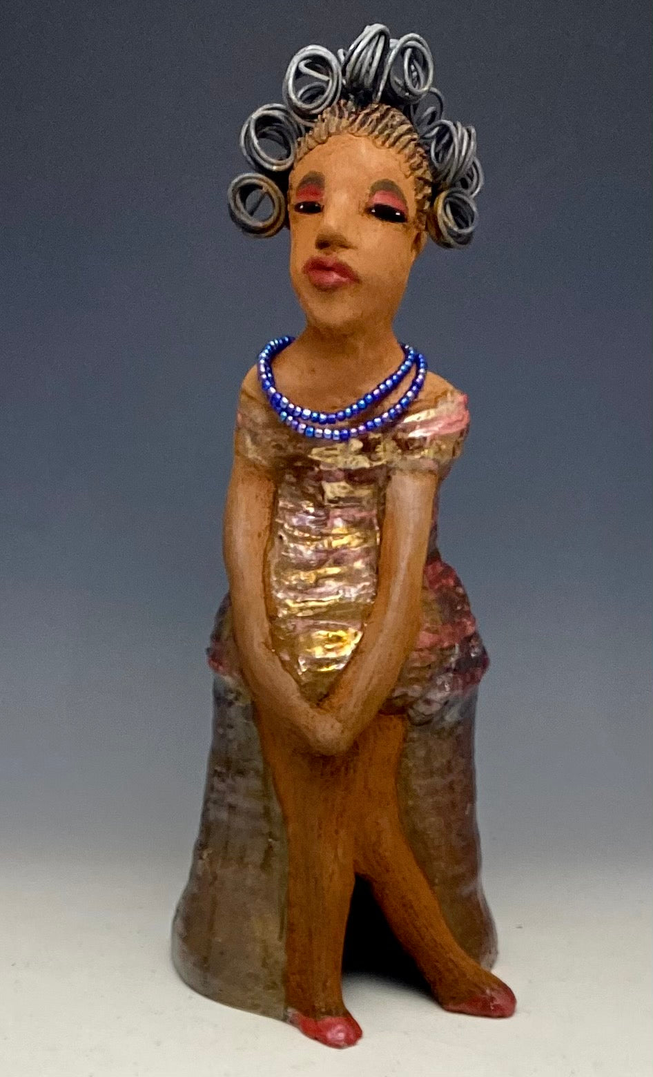 You know someone like Kim. One who reaps elegance and sophistication! Kim stands 13" x 5" x 7" and weighs 2.07 lbs. She has a lovely light beige complexion with over 4 feet of 16 gauge spiral coiled wire hair. Kim's metallic copper red and gold dress is accented by a multicolored aqua blue beaded necklace. Kim's maroon eye shadow gives her a distinguished mystical look. One must wonder what is Kim thinking? Kim sits with her legs crossed as she awaits a special place to grace your home.