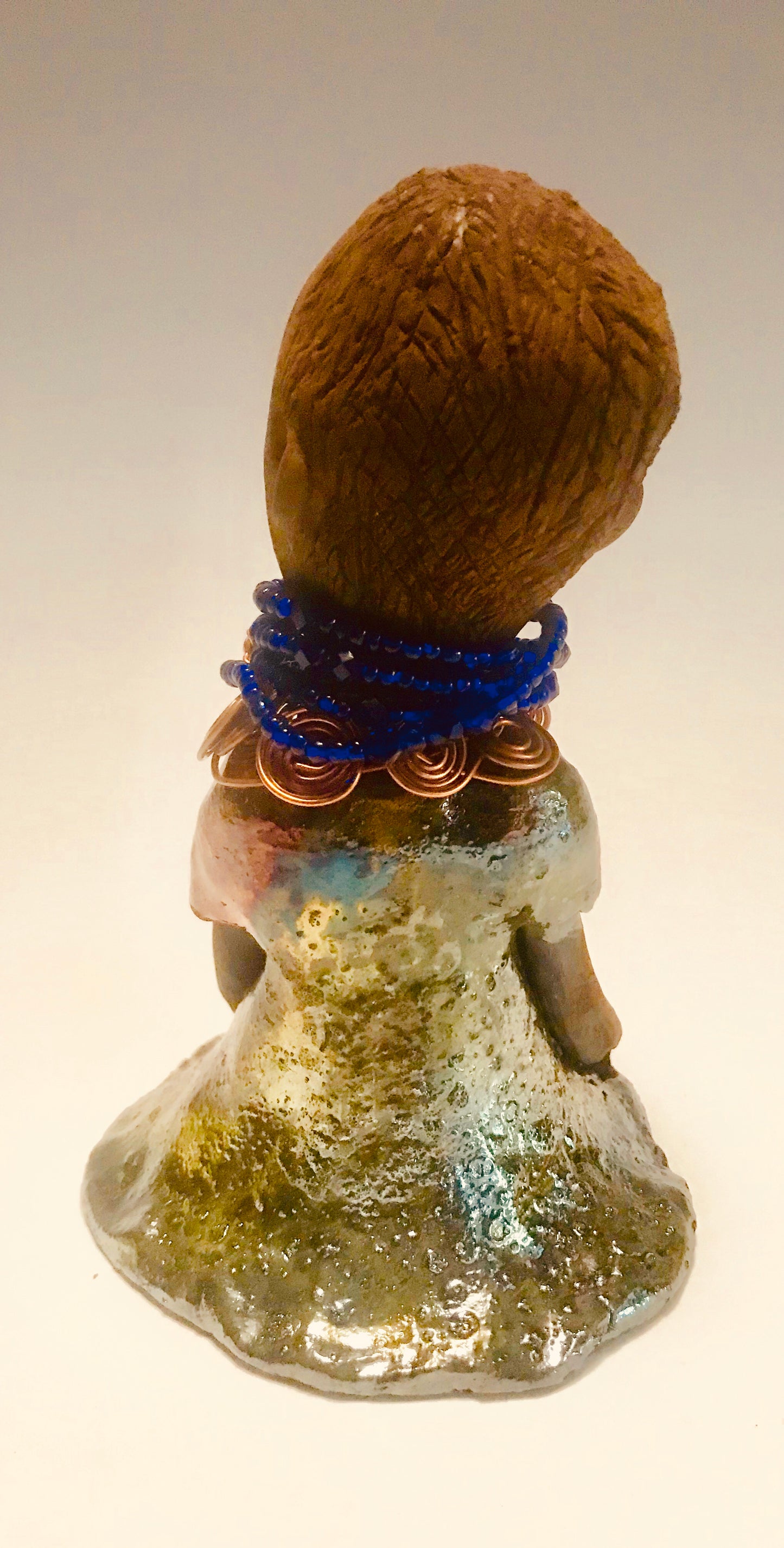 Sade stands 7.5" x 5" x 5" and weighs 1.04 lbs. She has a lovely honey brown complexion with reddish brown lips. She has a short braided hairstyle. Sade has a colorful metallic antique copper glazed dress. She wears spiral copper wire necklaces on top of an aqua blue beaded collar. Sade long loving arms rest at her side. With accent blue eye shadow and eyes wide opened, Sade has hopes of finding a new home.