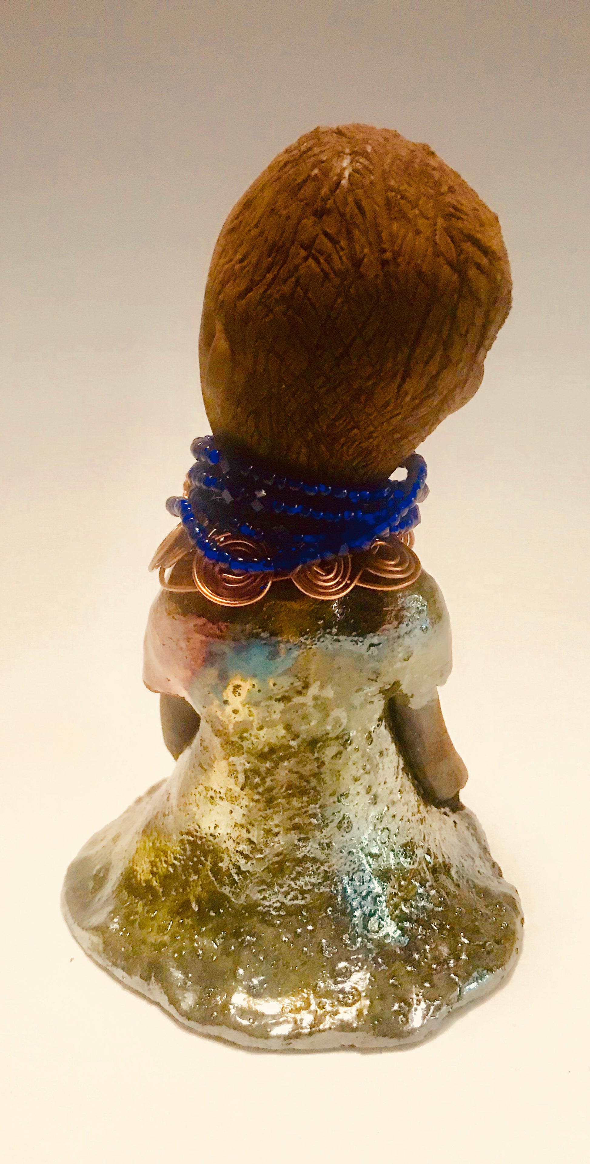 Sade stands 7.5" x 5" x 5" and weighs 1.04 lbs. She has a lovely honey brown complexion with reddish brown lips. She has a short braided hairstyle.  Sade  has a colorful metallic antique copper glazed dress. She wears spiral copper wire necklaces on top of an aqua blue beaded collar. Sade long loving arms rest at her side. With accent blue eye shadow and eyes wide opened, Sade has hopes of finding a new home.