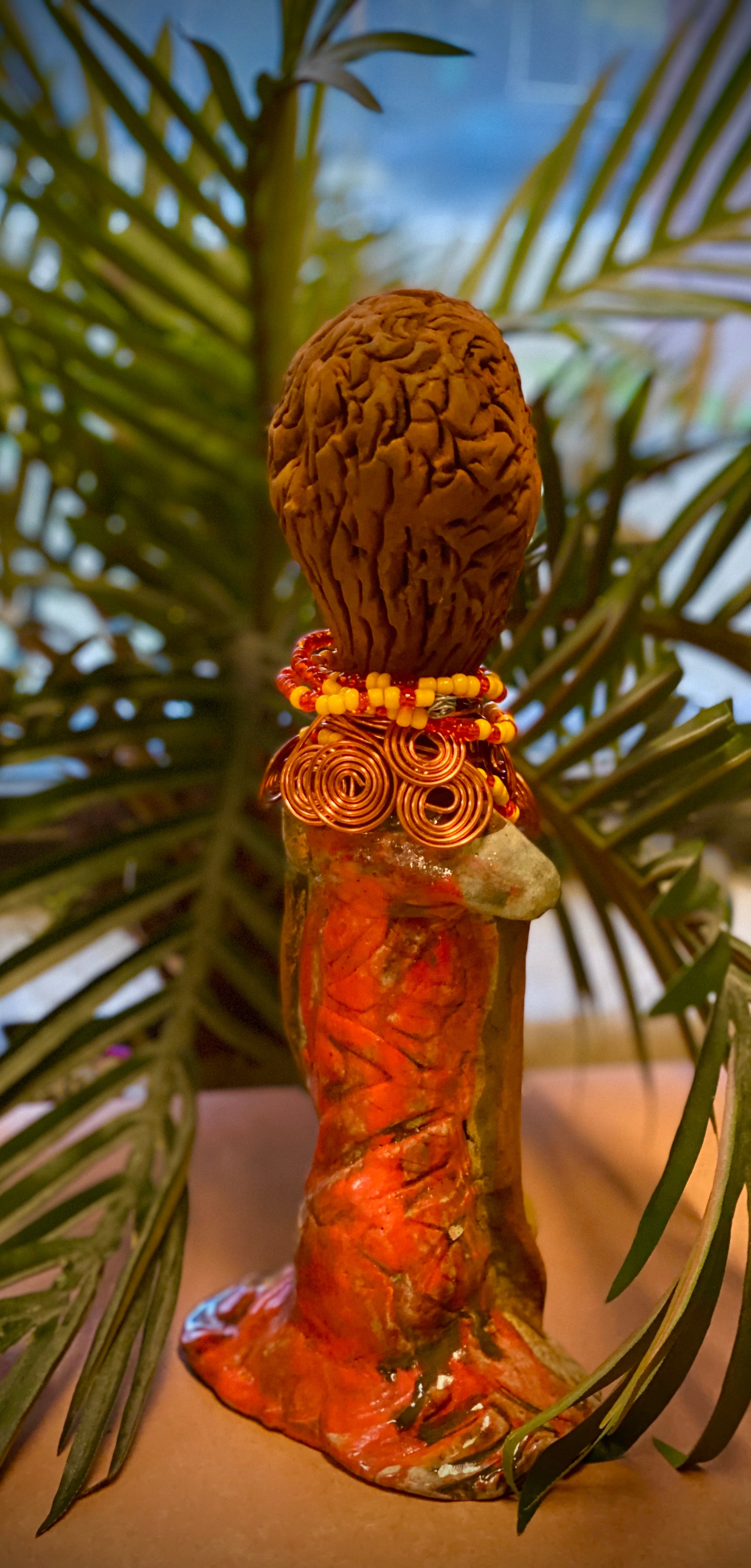 Reandra has a colorful reddish metallic green antique copper glazed dress. She wears a spiral copper wire necklace on top of a yellow red beaded collar. With eyes slightly opened, Reandra has hopes of finding a new home.
