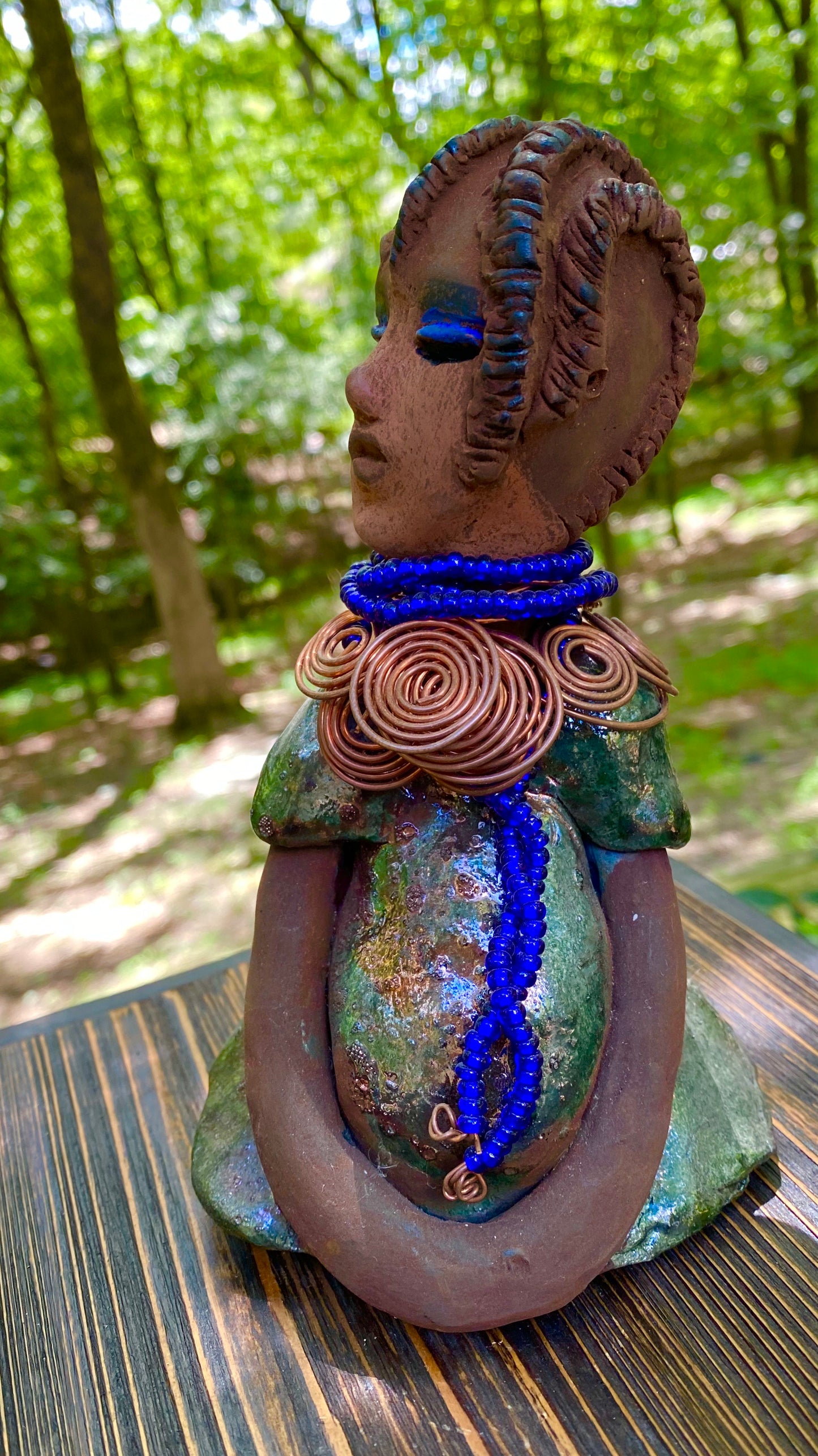 Meet Ife! Ife stands 7.5" x 4" x 5" and weighs 1.02 lbs. She has a lovely honey brown complexion with reddish brown lips. She has a short braided hairstyle. Ife has a colorful metallic antique copper glazed dress. She wears spiral copper wire necklaces on top of an aqua blue beaded collar. Ife long loving arms rest at her side. With accent blue eye shadow and eyes wide opened, Ife has hopes of finding a new home.