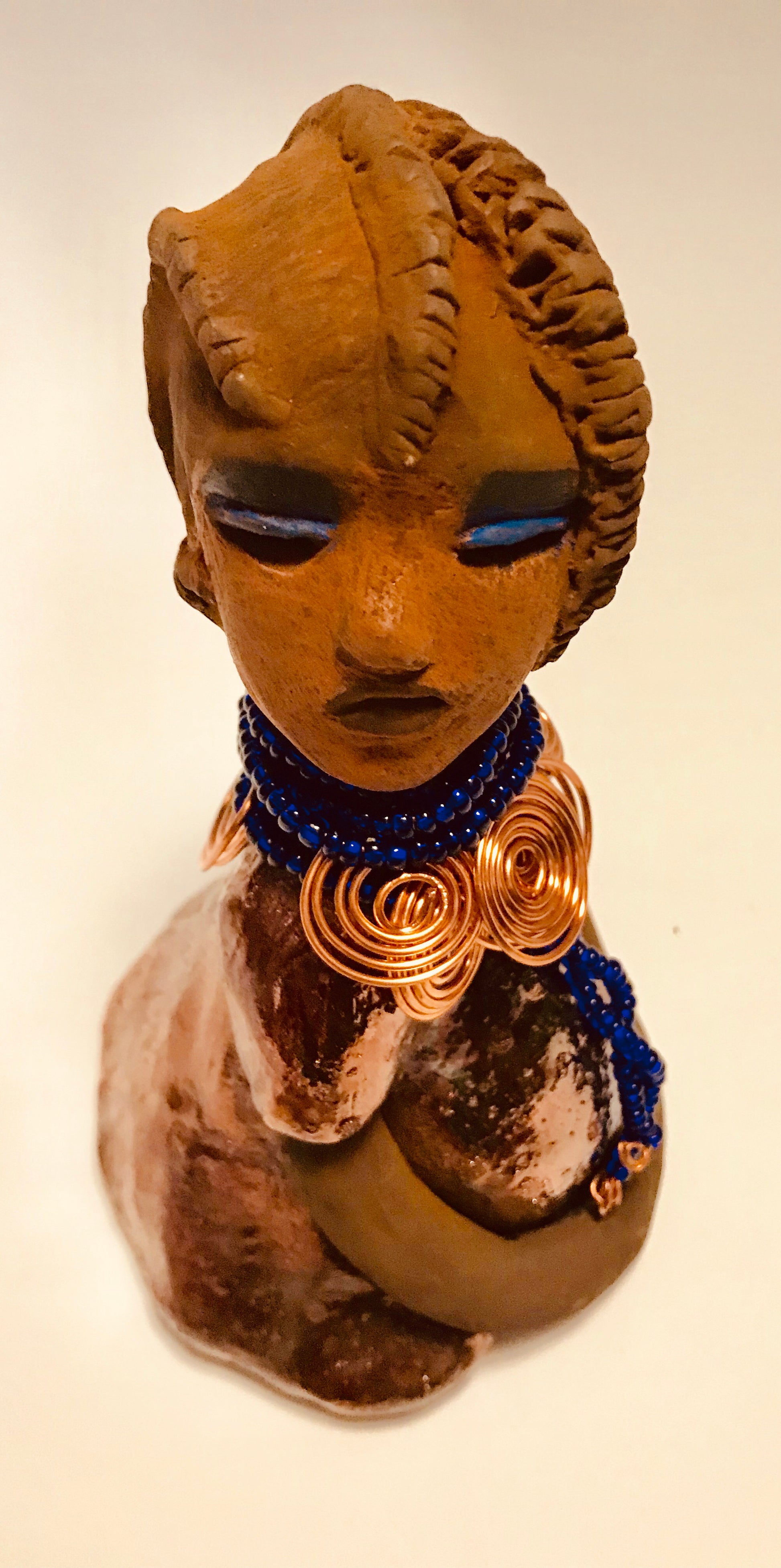 Ife stands 7.5" x 4" x 5" and weighs 1.02  lbs. She has a lovely honey brown complexion with reddish brown lips. She has a short braided hairstyle.      Ife has a colorful metallic antique copper glazed dress.     She wears spiral copper wire necklaces on top of an aqua blue beaded collar.     Ife long loving arms rest at her side.     With accent blue eye shadow and eyes wide opened, Ife has hopes of finding a new home.