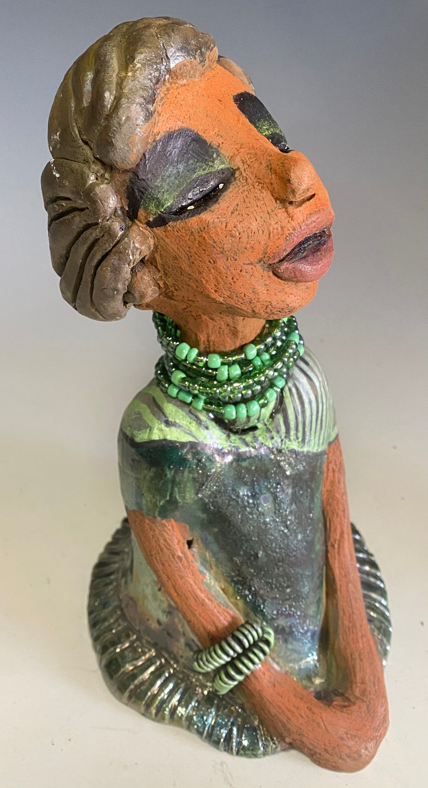 Meet Cora! Cora stands 9" x 5" x 6" and weighs 1.12 lbs. She has a lovely beige brown complexion with long etched braided clay hair. Cora’s long loving arms rest beside her copper green dress. Her dress is alligator green with a green coiled arm band. Cora wears a long string mixed green beaded necklace. Cora is a sophisticated lady that will grace your home.