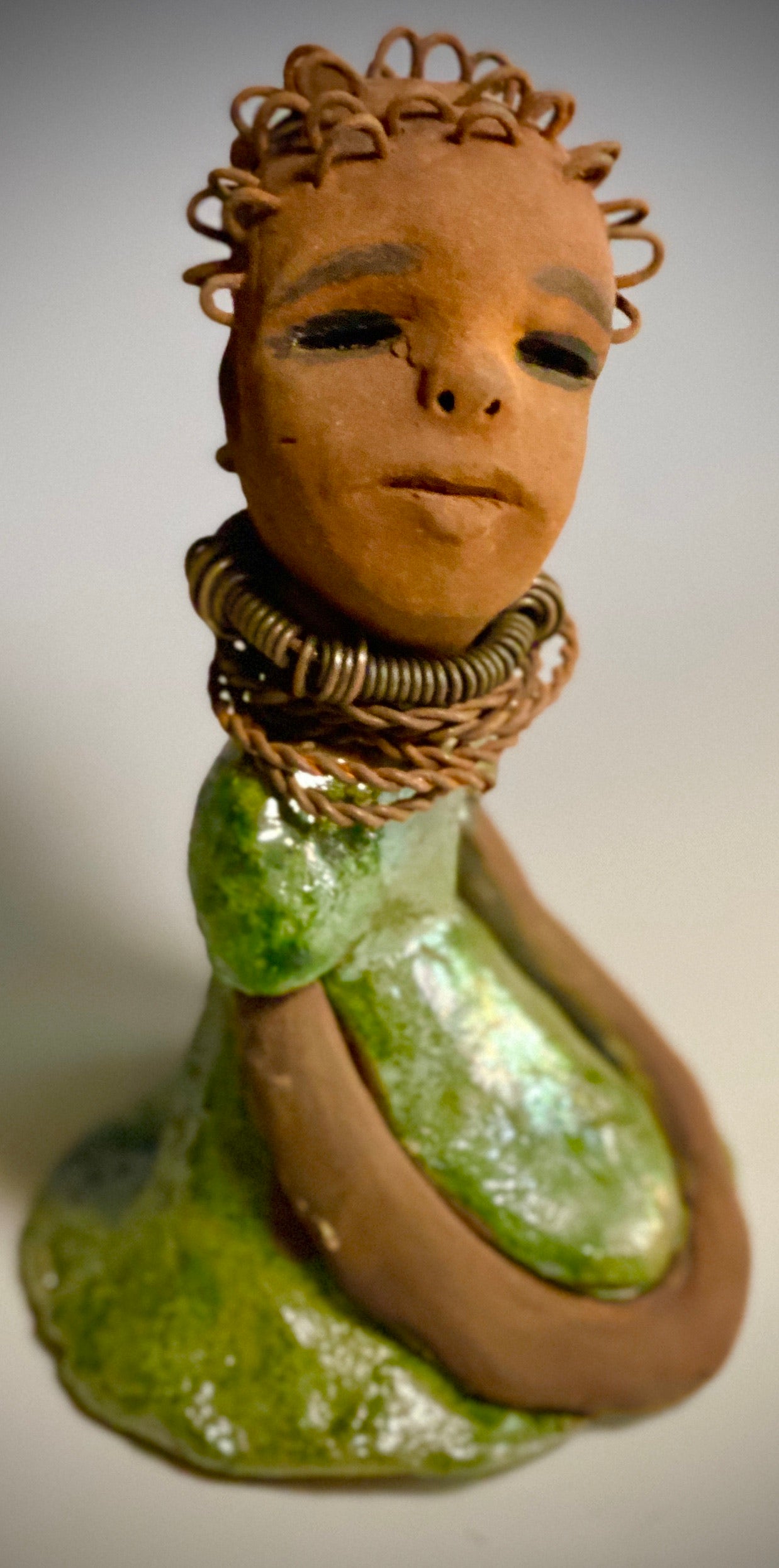Meet Nura! Nura stands 7.5" x 5" x 5" and weighs 1.06 lbs. She has a lovely honey brown complexion with reddish brown lips. She has a wire braided hairstyle. Nura has a colorful metallic green antique copper glazed dress. She wears spiral wire necklaces With her eyes wide opened, Nura has hopes of finding her way into your home.