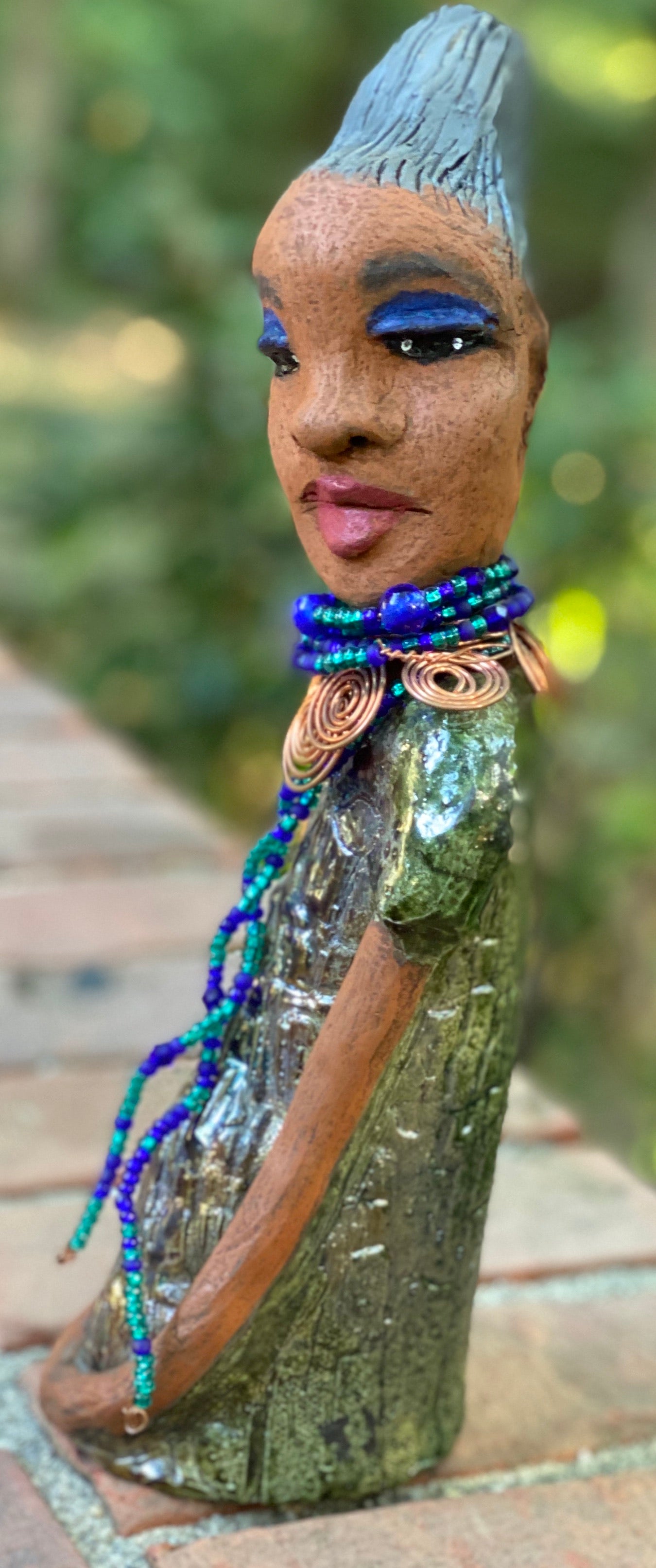 Meet Emma Again! Emma went and got her a makeover too. She was jealous of Eonna! Emma stands 12.5" x 3. 5" x 4" and weighs 2.04 lbs. She has a lovely honey brown complexion with reddish brown lips. She has a braided clay hairstyle. Emma has a colorful metallic green antique copper glazed dress. She wears a spiral copper wire necklace on top of a multicolored blue beaded collar. Check out Emma before and after pics. With eyes wide opened, Emma has hopes of finding her way into your home!