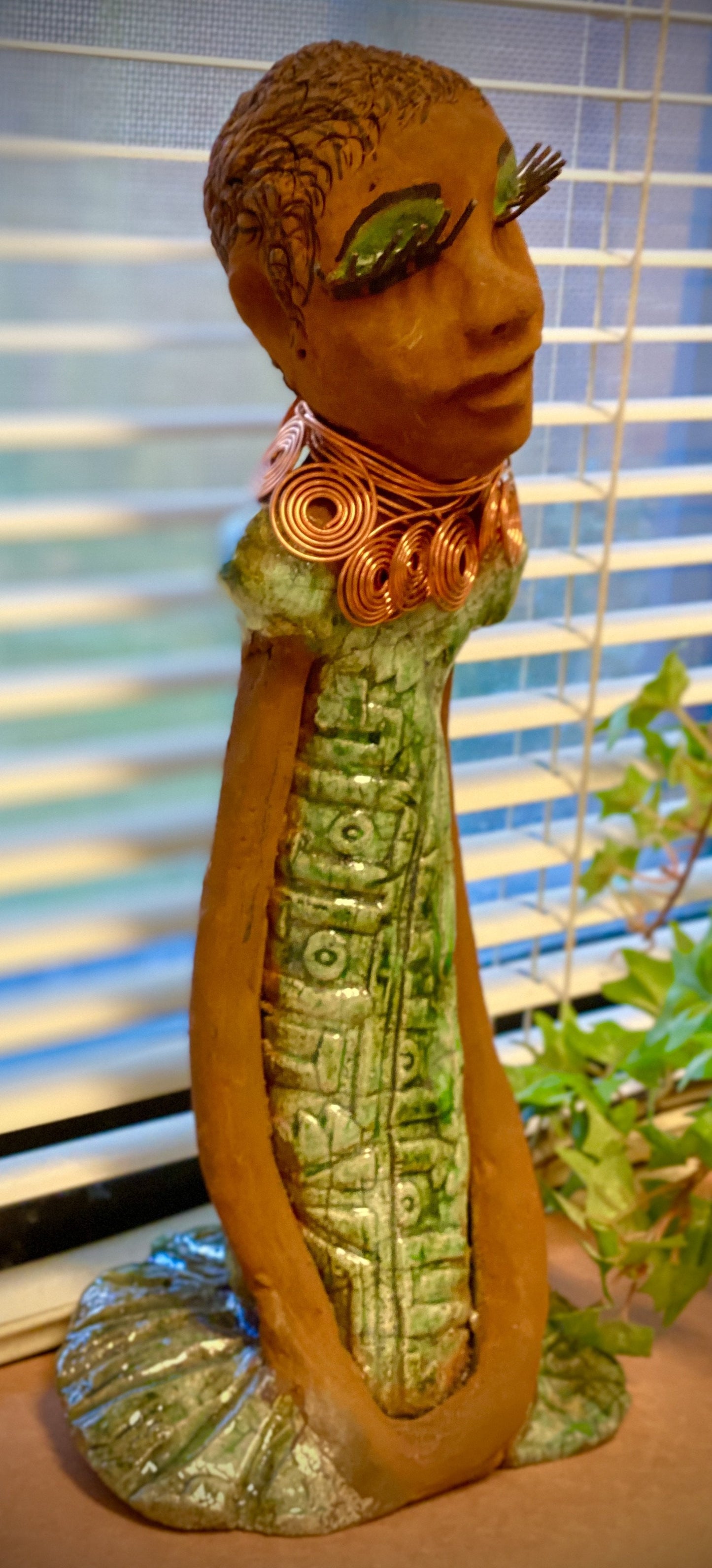 Gabrielle stands 14" x 6" x 5.5" and weighs 2.15 lbs. She has a lovely honey brown complexion with green eye shadow. Her hairstyle is etched in clay with tribal markings. Gabrielle long loving arms rest beside her alligator green dress. She wears a spiral copper necklace. Gabrielle is a sophisticated lady that will grace your home.