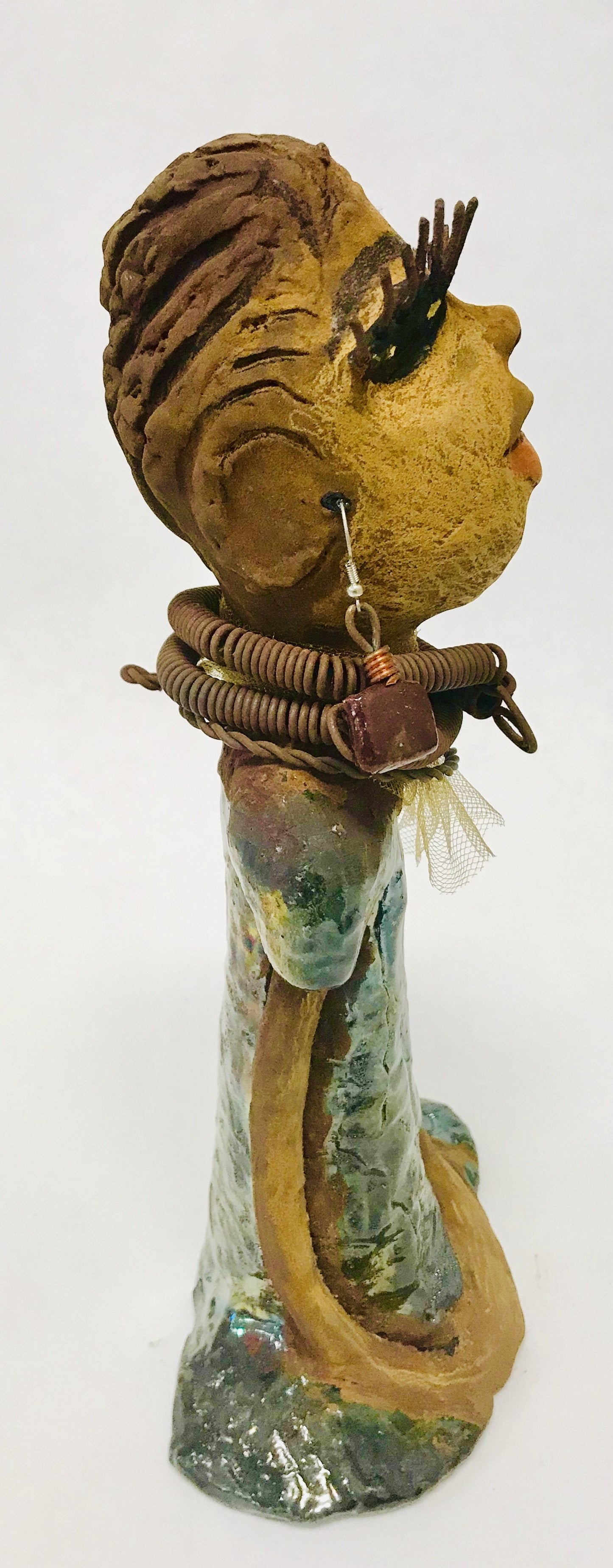 Catera stands 10" x 4" x 4" and weighs 2.7 lbs. She has a lovely honey brown complexion with reddish brown lips. She has a braided hairstyle. Catera has a colorful metallic green antique copper glazed dress. She wears a yellow bead necklace with raku beads. Her long loving arms are resting at her side. With long lashes and eyes wide opened, Catera has hopes of finding a new home.