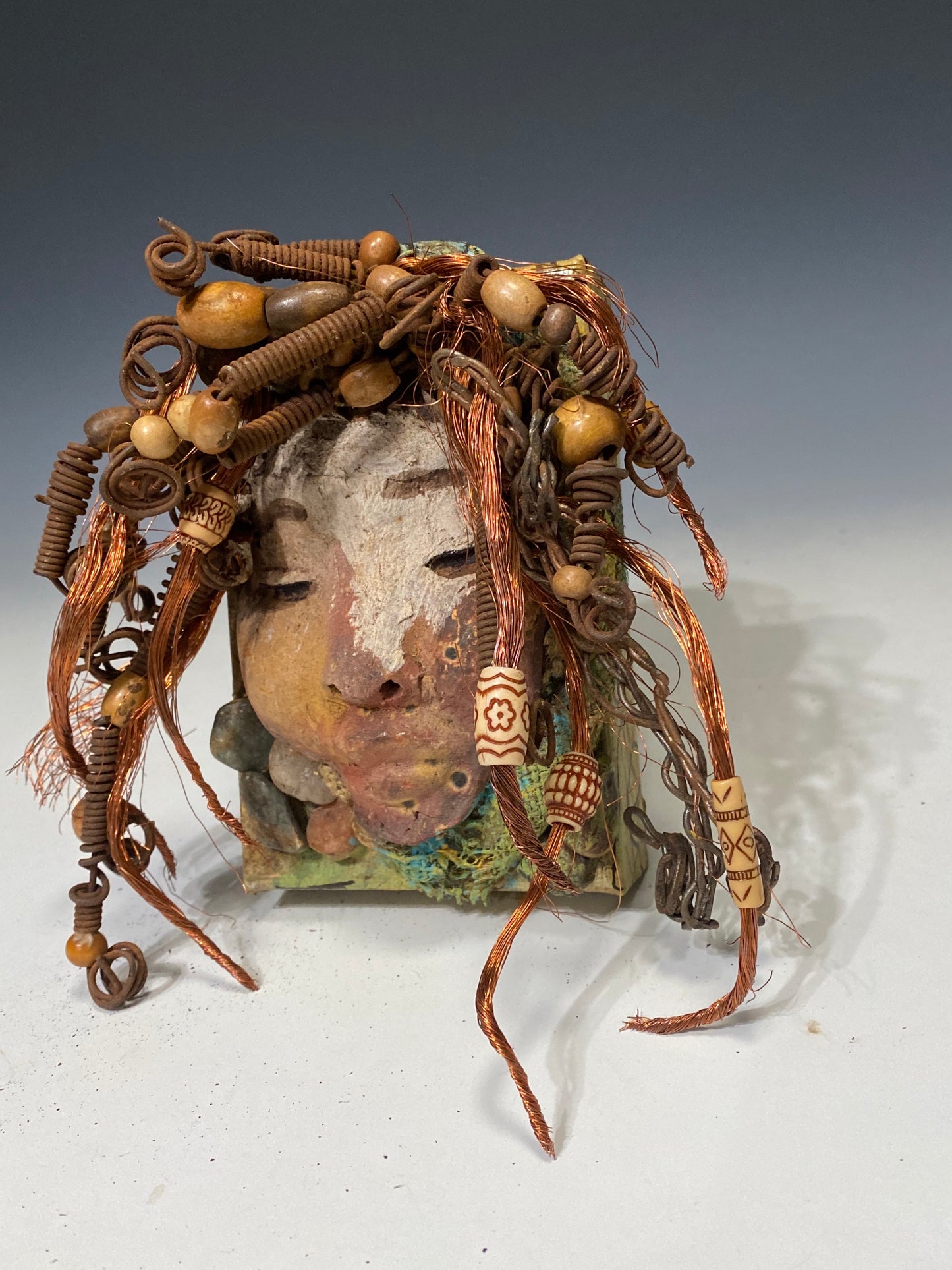 I started making art soon after seeing authentic African artwork at the Smithsonian Museum of African Art. I was in total awe. Lulu was inspired by my visit there. Lulu is uniquely mounted on a painted 6"x 4"x 2" canvas. It weighs 1.15 lbs. Her face has a two tone off white and earthy crackle glaze. Lulu has hand coiled wire, white and multi color beads, textured earthy cloth..