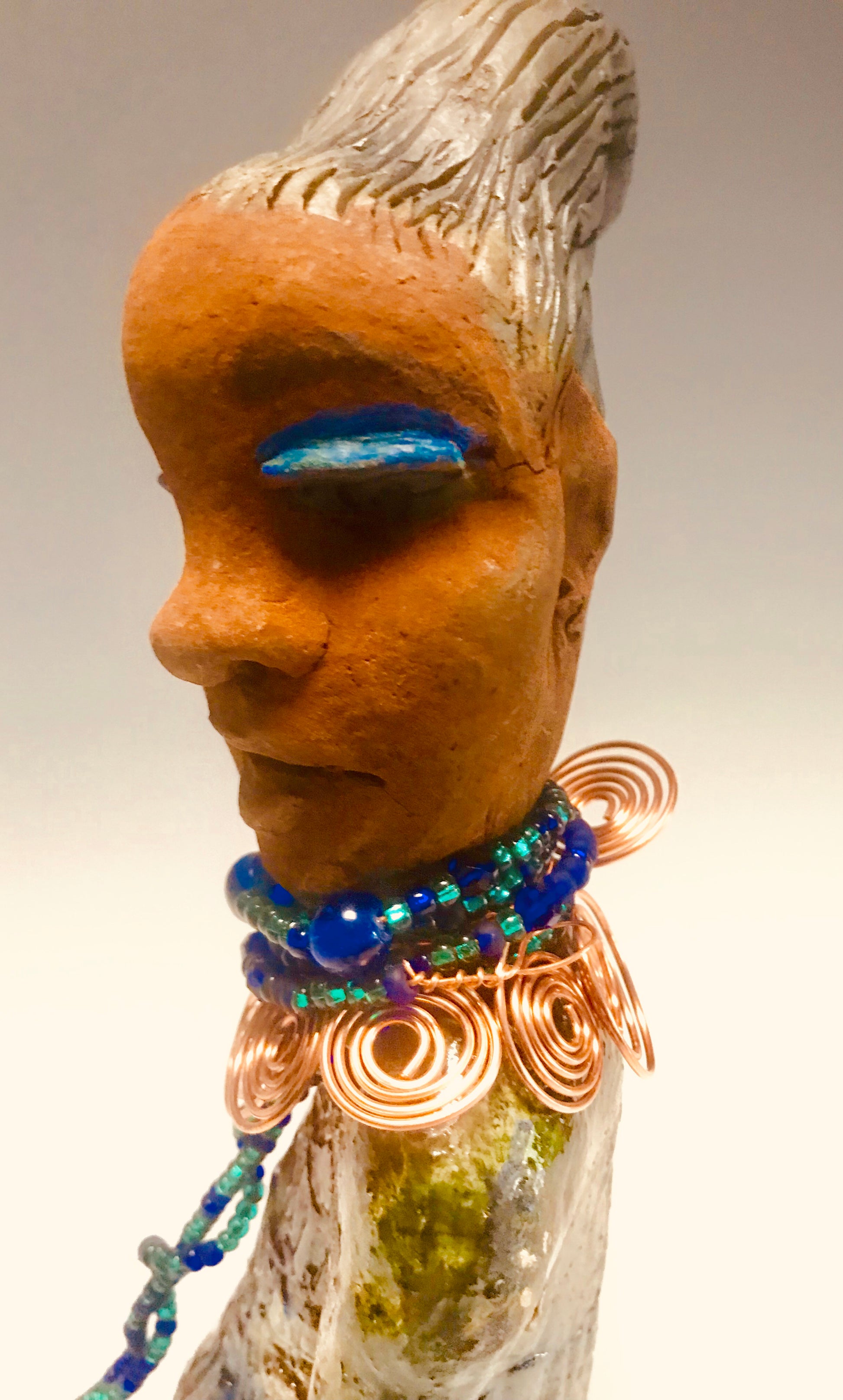 Emma stands 12.5" x3. 5" x 4" and weighs 2.04 lbs. She has a lovely honey brown complexion with  reddish brown lips. She has a braided hairstyle.  Emma has a colorful metallic green antique copper glazed dress. She wears a spiral copper wire necklace on top of a multicolored blue beaded collar. With eyes wide opened,  Emma has hopes of finding a new home.