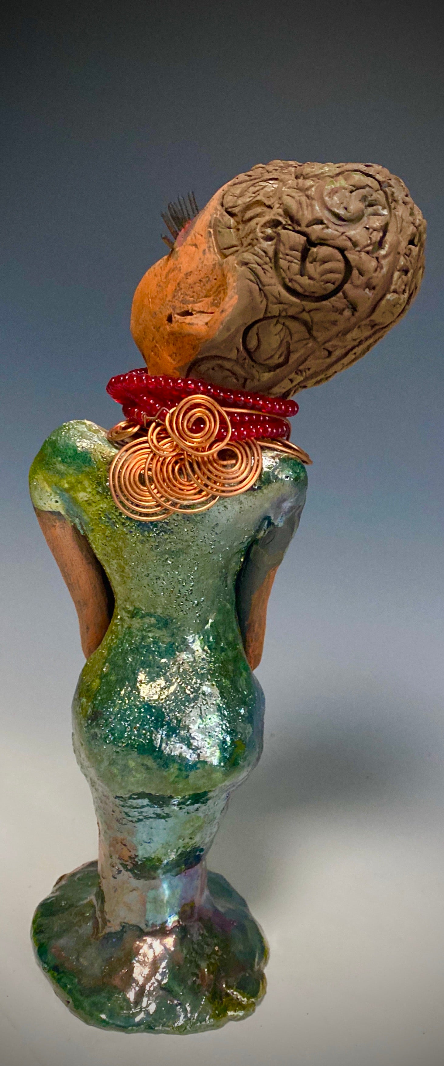 Meet Mona! Mona stands 11.5" x 4" x 4" and weighs 1.81 lbs. She has a lovely chocolate brown complexion with chocolate brown lips. She has a braided hairstyle.  Mona has a colorful metallic antique copper glazed dress. She wears a spiral copper wire necklaces on top of a beaded ruby red collar. With long lashes and eyes wide opened, Mona has hopes of finding a new home. You may have noticed, Mona is the one of a few herdew  sculptures that wears a face  covering during this unprecedented time.