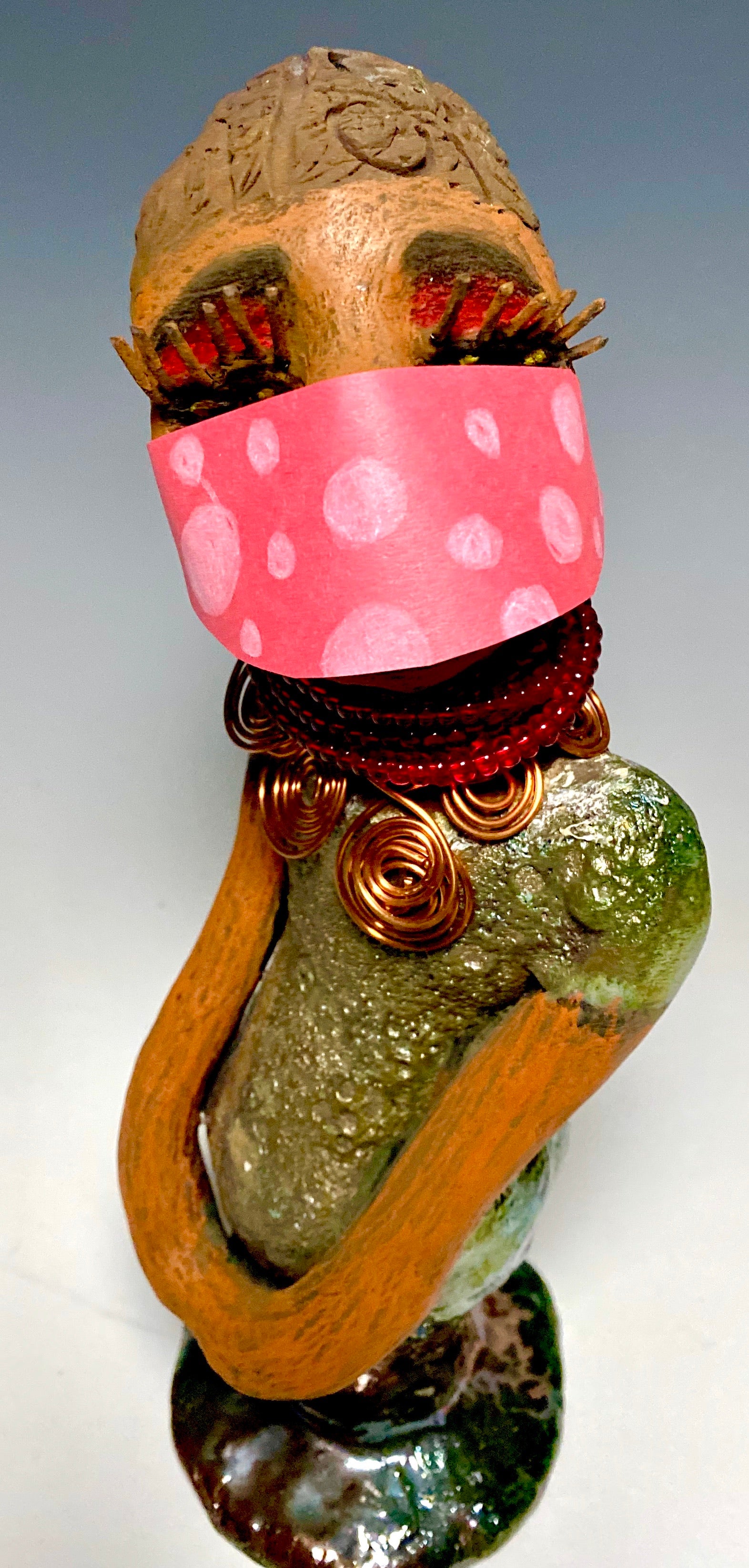 Meet Mona! Mona stands 11.5" x 4" x 4" and weighs 1.81 lbs. She has a lovely chocolate brown complexion with chocolate brown lips. She has a braided hairstyle.  Mona has a colorful metallic antique copper glazed dress. She wears a spiral copper wire necklaces on top of a beaded ruby red collar. With long lashes and eyes wide opened, Mona has hopes of finding a new home. You may have noticed, Mona is the one of a few herdew  sculptures that wears a face mask during this unprecedented time.