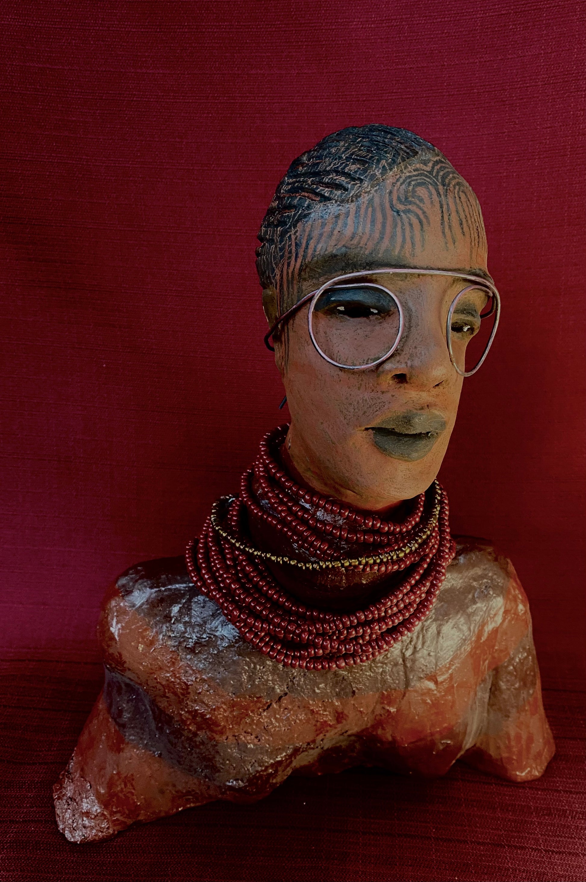 Meet Harriet! Harriet stands 13" x 11" x 5" and weighs 5 lbs. She wears a coral striped dress with a string of ruby red and gold beads. Harriet has a honey brown rust complexion  and distinct eyes. Her eyes  are slightly opened underneath her glasses... She has mixed black and copper streaks of straight corn rolls of clay hair. She has a straight look of concern as to what is about to take place.