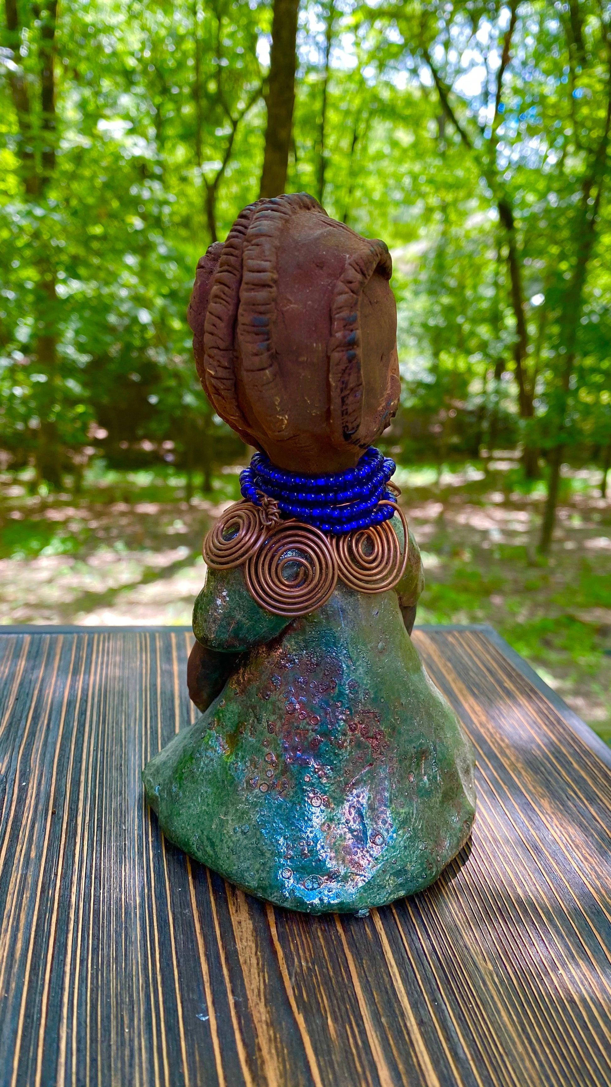Meet  Ife! Ife stands 7.5" x 4" x 5" and weighs 1.02  lbs. She has a lovely honey brown complexion with reddish brown lips. She has a short braided hairstyle.  Ife has a colorful metallic antique copper glazed dress. She wears spiral copper wire necklaces on top of an aqua blue beaded collar. Ife long loving arms rest at her side. With accent blue eye shadow and eyes wide opened, Ife has hopes of finding a new home.
