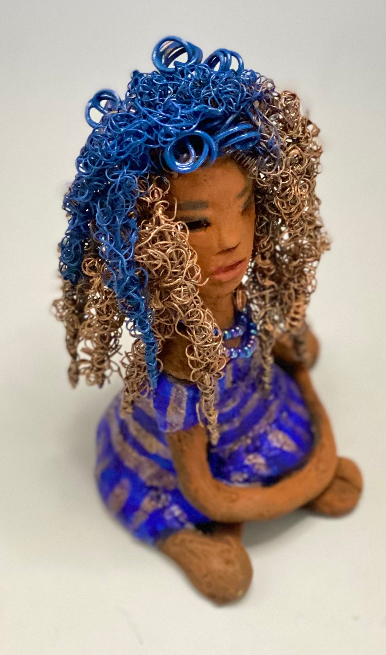 Sherrie stands 6" x 4" x 4.5" and weighs 12.6 ozs. She has a lovely honey brown complexion with reddish brown lips. Sherrie's hairstyle is made up of over 75 feet of twisted 24 gauge wire. Sherrie has a striped metallic copper and blue glazed dress. She wears a aqua blue beaded necklaces. With eyes wide opened, Sherrie has hope of finding a new home. She will make an excellent starter piece from the HerDew collection!