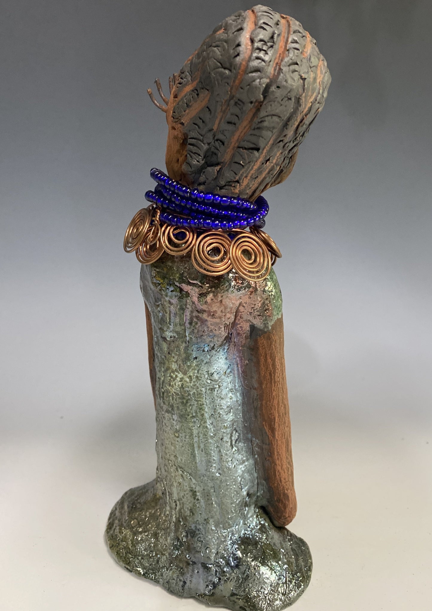 Meet Ophelia! Ophelia stands 7.5" x 4" x 5" and weighs 1.02 lbs. She has a lovely honey brown complexion with reddish brown lips. She has a short braided hairstyle. Ophelia has a colorful metallic antique copper glazed dress. She wears spiral copper wire necklaces on top of an aqua blue beaded collar. Ophelia long loving arms rest at her side. With accent blue eye shadow and eyes wide opened, Ophelia has hopes of finding her way into your home!