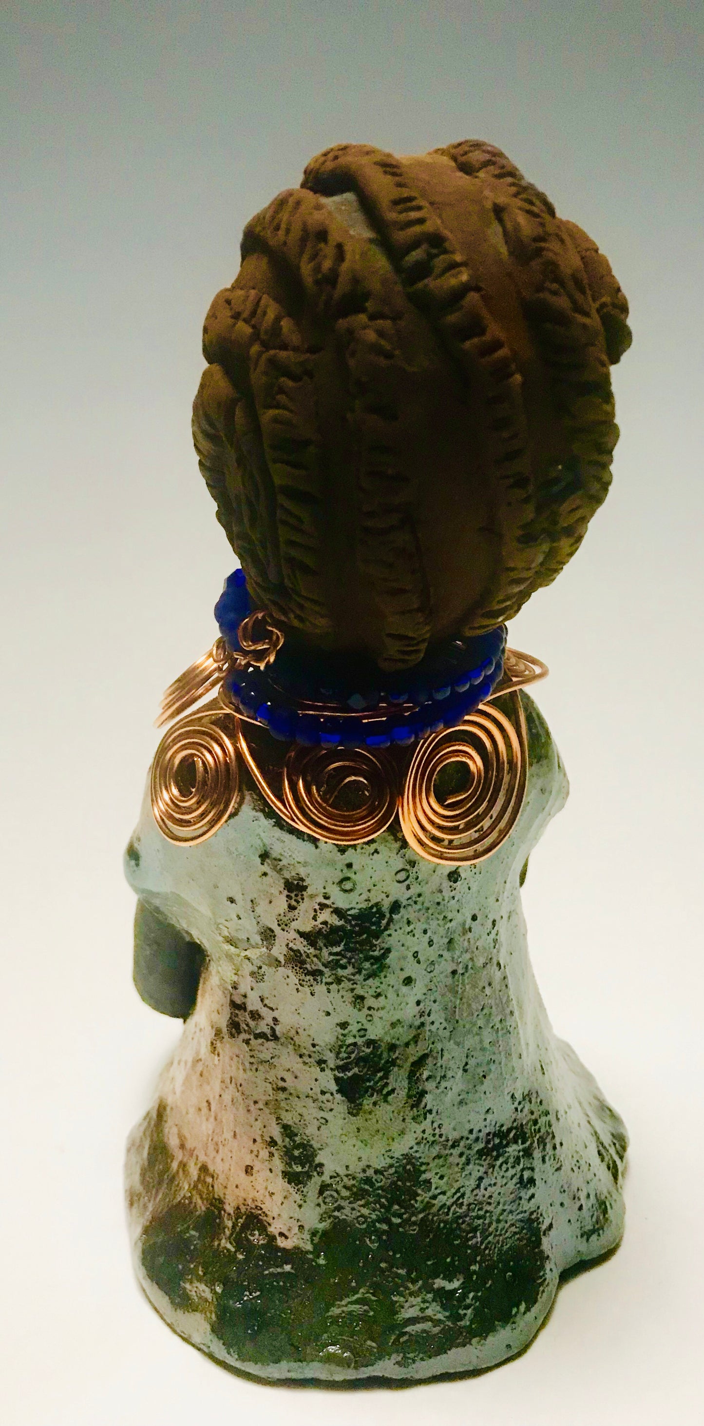 Anja stands 7" x 4" x 4" and weighs 1.01 lbs. She has a lovely honey brown complexion with reddish brown lips. She has a braided hairstyle. Anja has a colorful metallic green antique copper glazed dress. She wears a spiral copper wire necklace on top of a blue beaded collar. With eyes slightly opened, Anja has hopes of finding a new home.