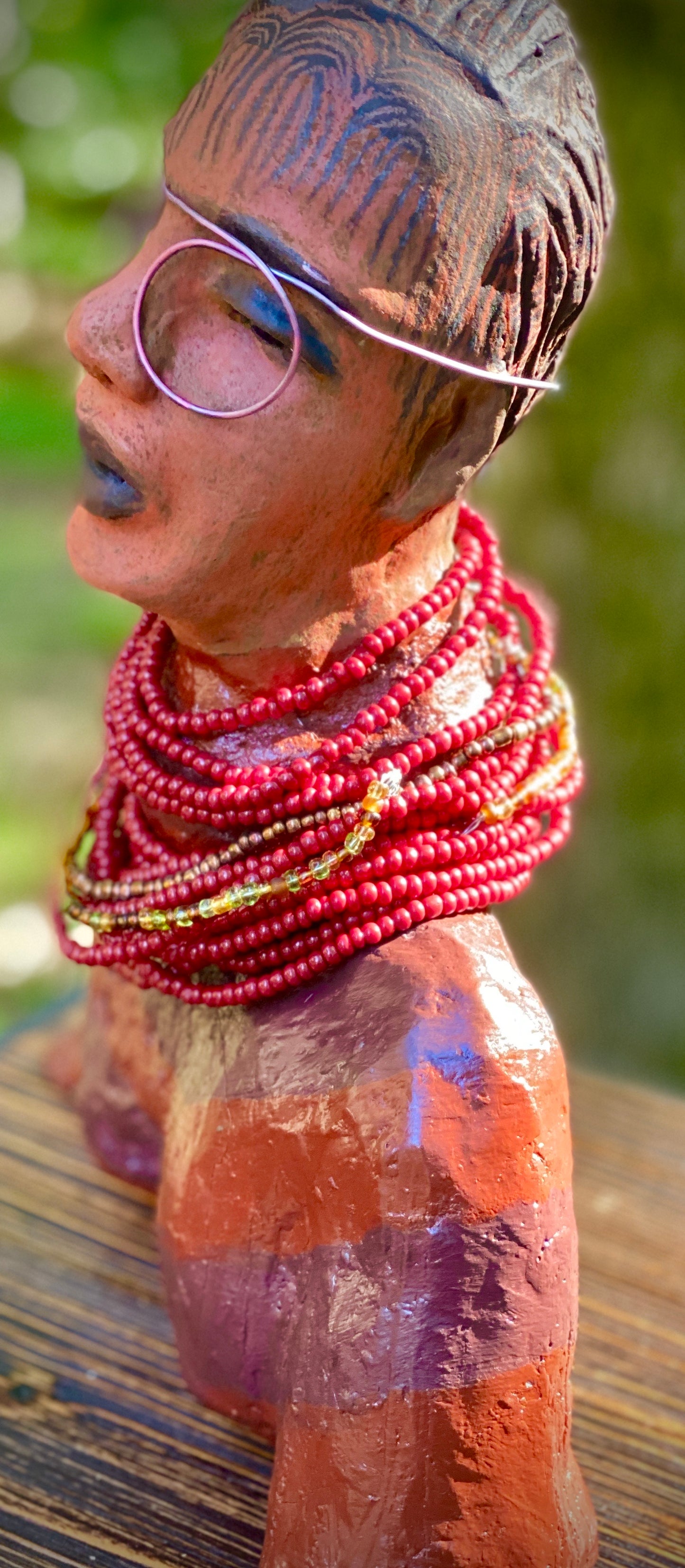 Meet Harriet! Harriet stands 13" x 11" x 5" and weighs 5 lbs. She wears a coral striped dress with a string of ruby red and gold beads. Harriet has a honey brown rust complexion and distinct eyes. Her eyes are slightly opened underneath her glasses... She has mixed black and copper streaks of straight corn rolls of clay hair. She has a straight look of concern as to what is about to take place.
