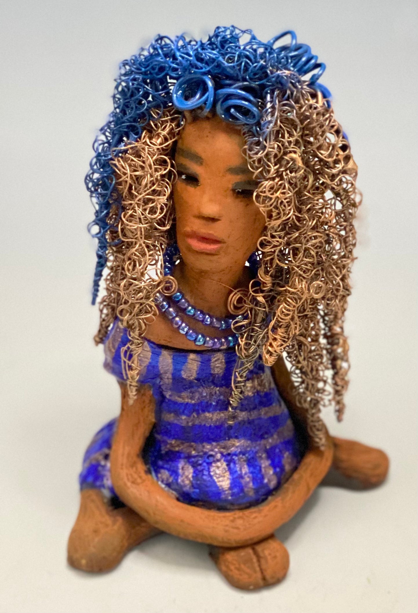 Sherrie stands 6" x 4" x 4.5" and weighs 12.6 ozs. She has a lovely honey brown complexion with reddish brown lips. Sherrie's hairstyle is made up of over 75 feet of twisted 24 gauge wire. Sherrie has a striped metallic copper and blue glazed dress. She wears a aqua blue beaded necklaces. With eyes wide opened, Sherrie has hope of finding a new home. She will make an excellent starter piece from the HerDew collection!
