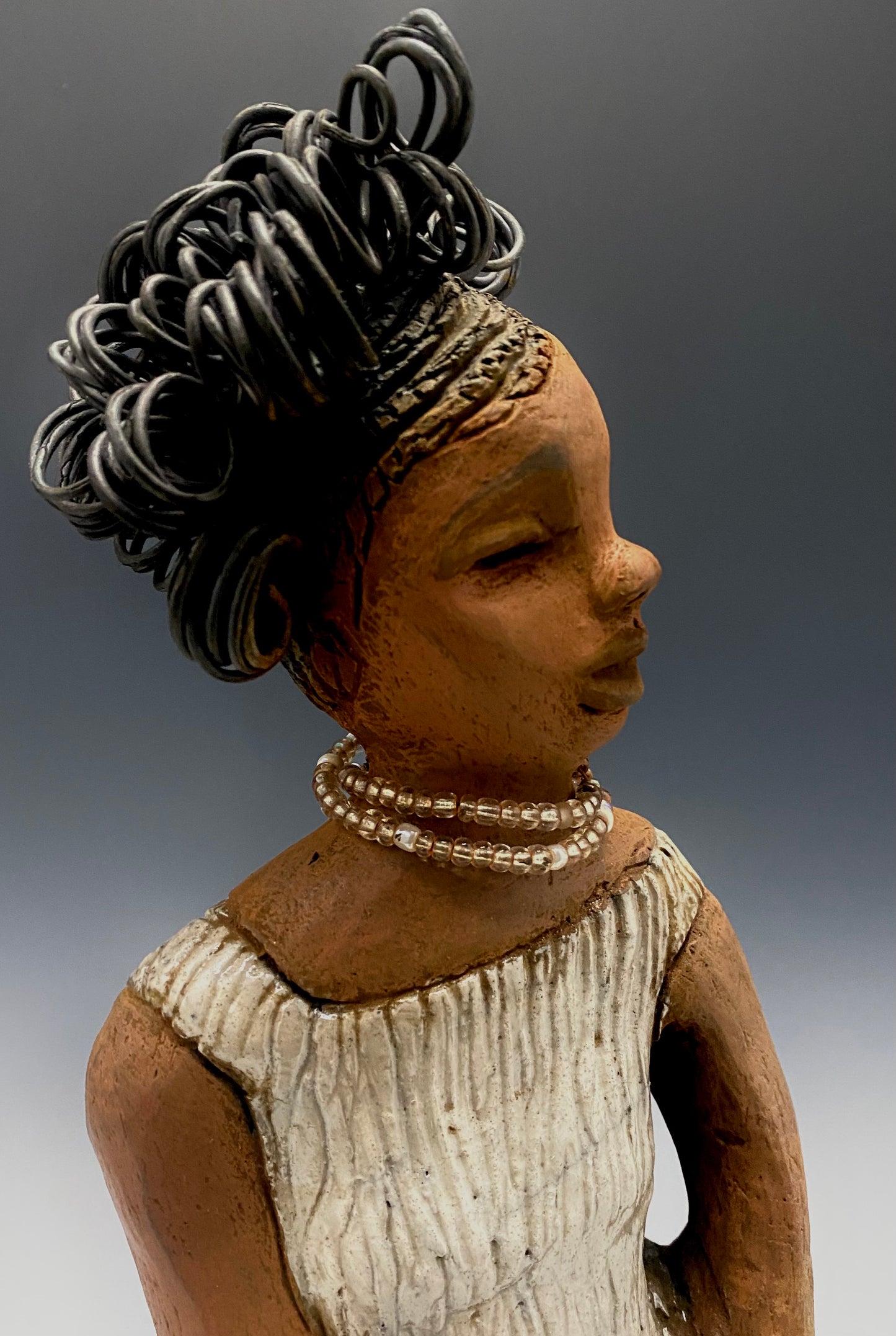 Meet Gwen! You know someone like Gwen. One who reaps elegance and sophistication! Gwen stands 13 " x 6" x 6" and weighs 3.07 lbs. She has a lovely brown complexion with over 5 feet of 16 gauge spiral coiled wire hair. Gwen's metallic off white dress is accented by a multicolored white and amber beaded necklace. Gwen’s straight face gives her a distinguished no nonsense look. Gwen sits with her hands rested as she awaits a special place to grace your home.