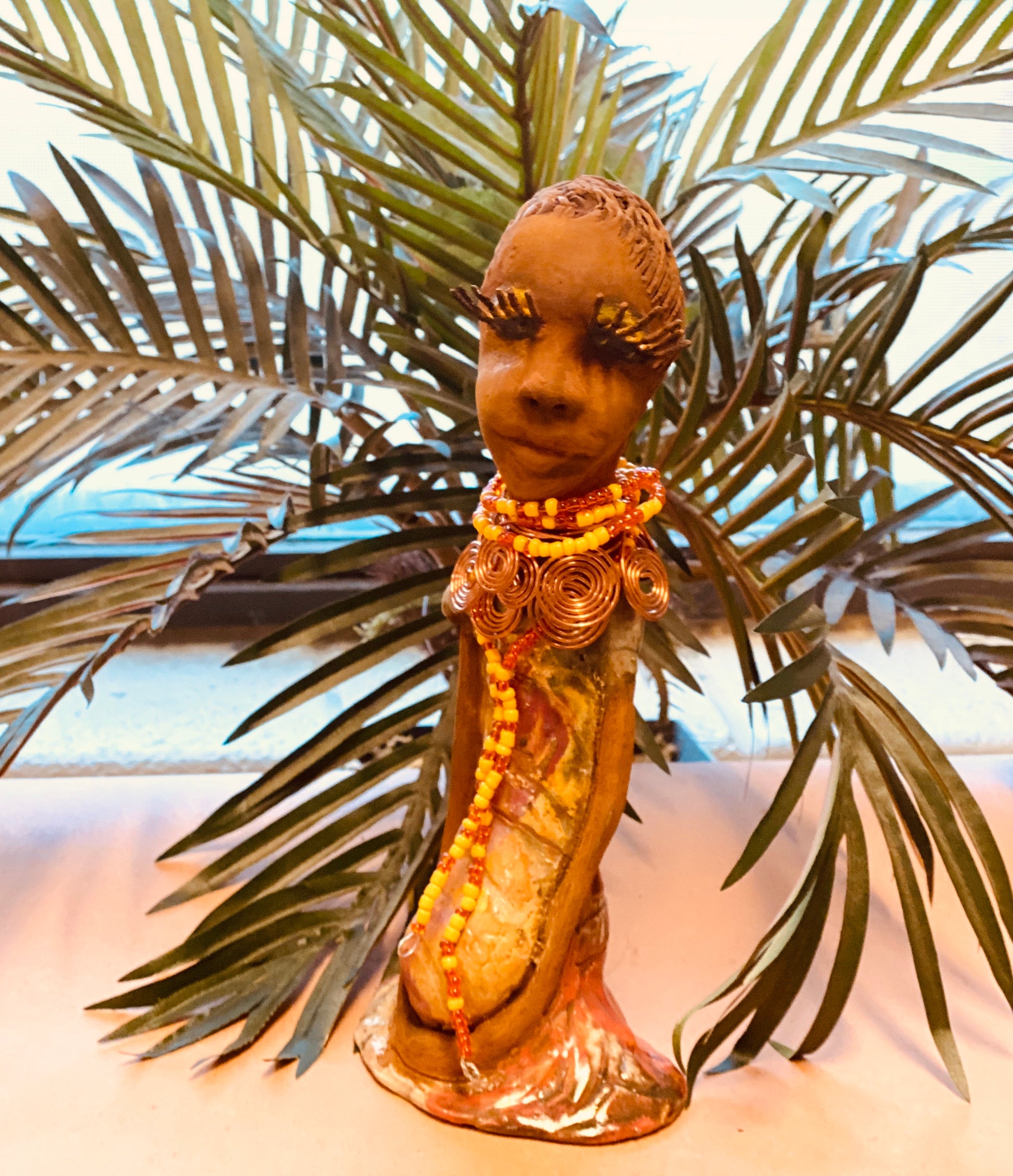 Reandra has a colorful reddish metallic green antique copper glazed dress. She wears a spiral copper wire necklace on top of a yellow red beaded collar. With eyes slightly opened, Reandra has hopes of finding a new home.