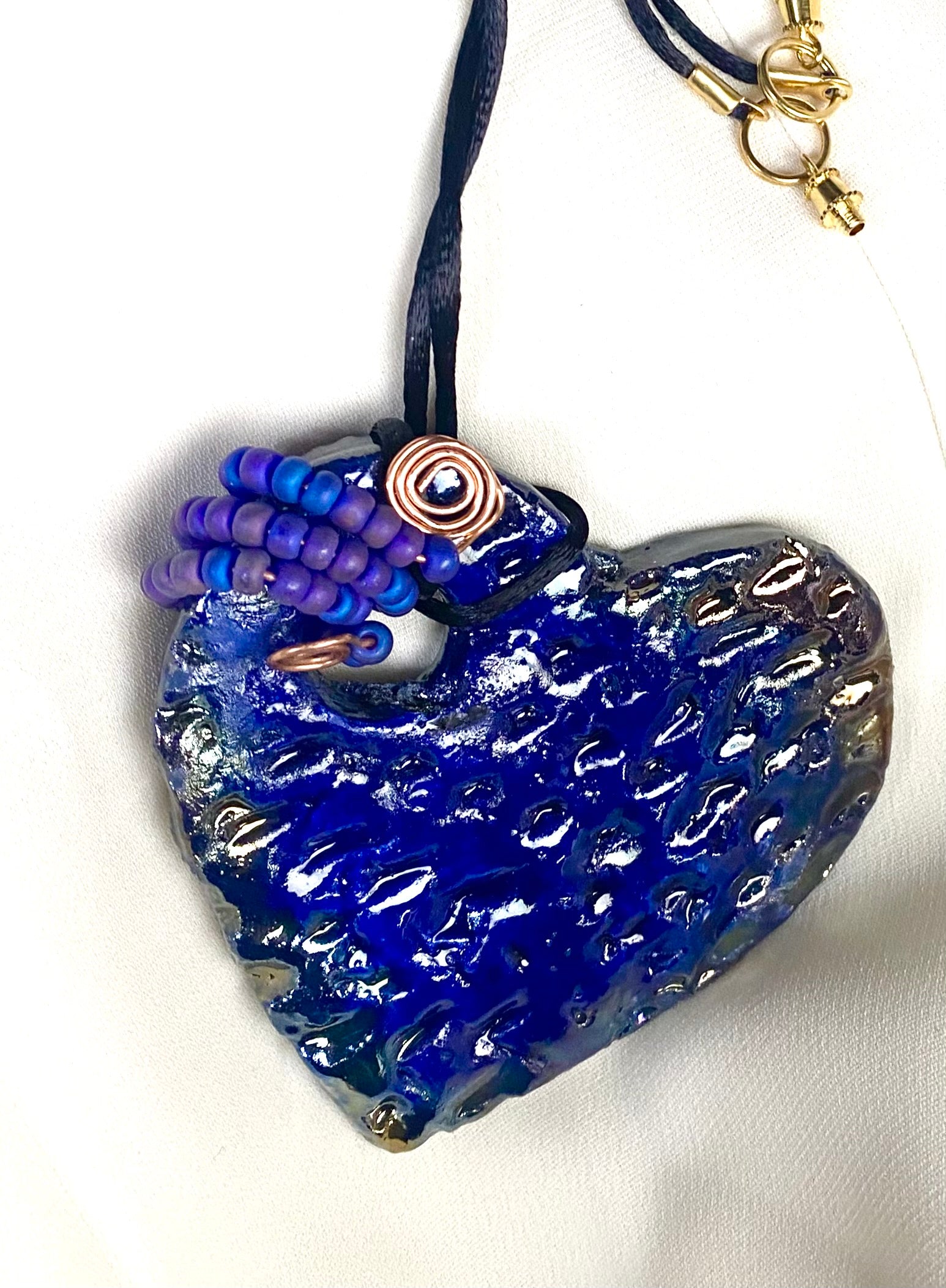 Have A Heart ! Each heart pendant is handmade with love! It is 3"x 3" and weighs approx. 3ozs. This pendant has a royal blue and gold metallic raku glazes that renders a unique translucent  patina. The heart  has a textured  pattern. It holds a spiral of off blue violet mini beads on a spiral copper wire. This pendant has a nice 12" black suede cord!