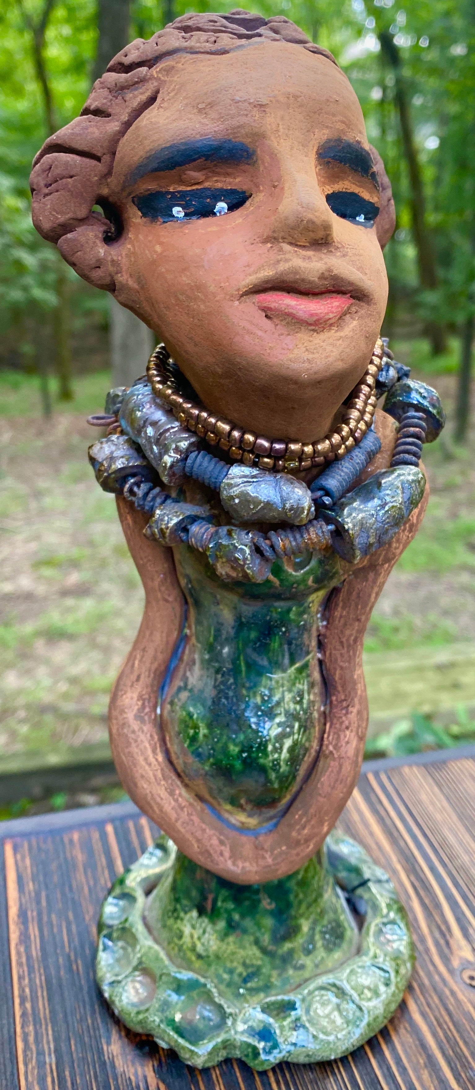Greta! Greta stands 11" x 4" x 4" and weighs 2.5 lbs. She has a lovely honey brown complexion with  reddish brown lips. She has a braided hairstyle.  Gretaya has a colorful metallic green antique copper glazed dress. She wears two raku beaded necklaces. With long eyes slightly opened, Greta has hopes of finding a new home.