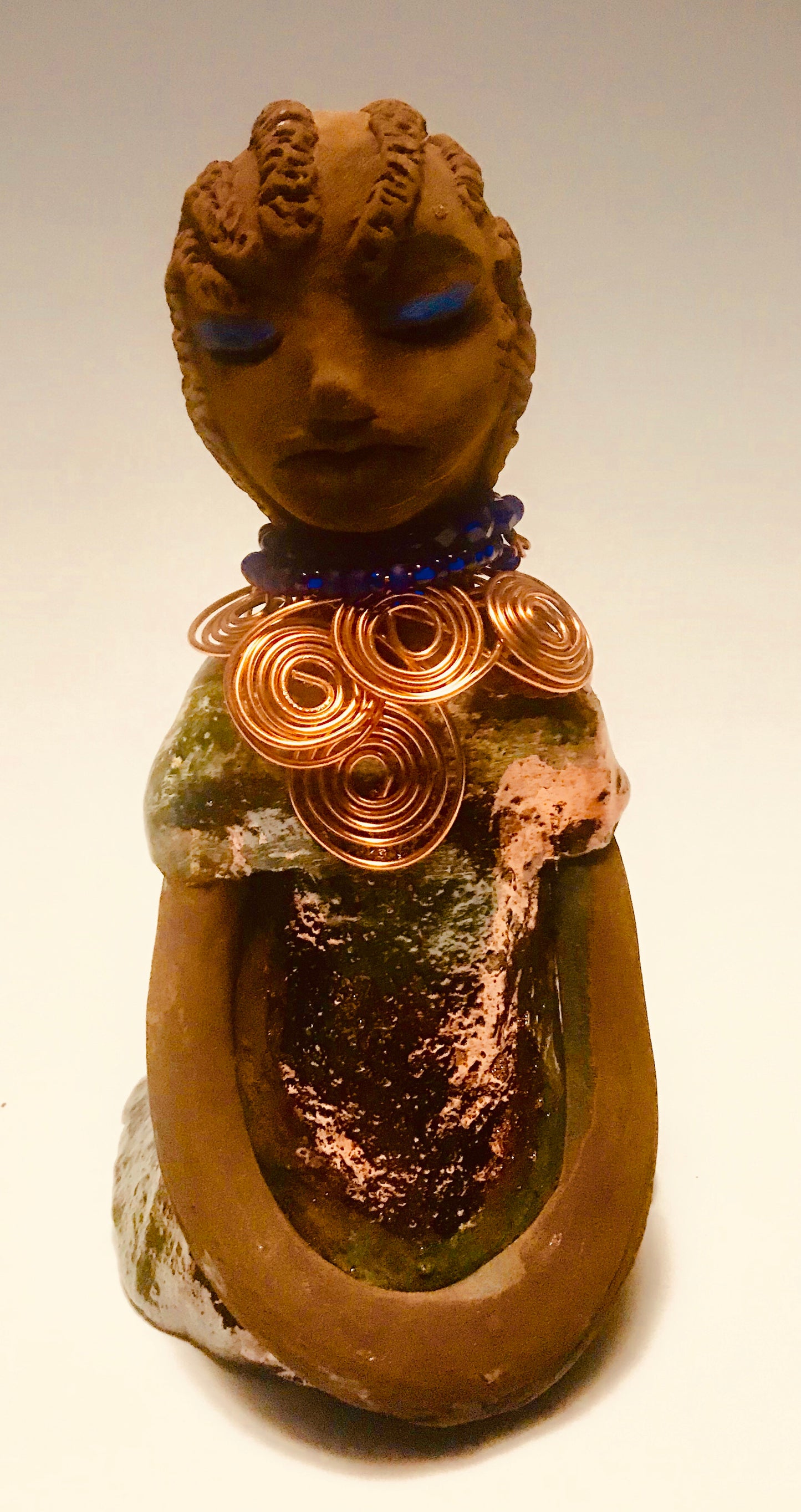 Anja stands 7" x 4" x 4" and weighs 1.01 lbs. She has a lovely honey brown complexion with reddish brown lips. She has a braided hairstyle. Anja has a colorful metallic green antique copper glazed dress. She wears a spiral copper wire necklace on top of a blue beaded collar. With eyes slightly opened, Anja has hopes of finding a new home.