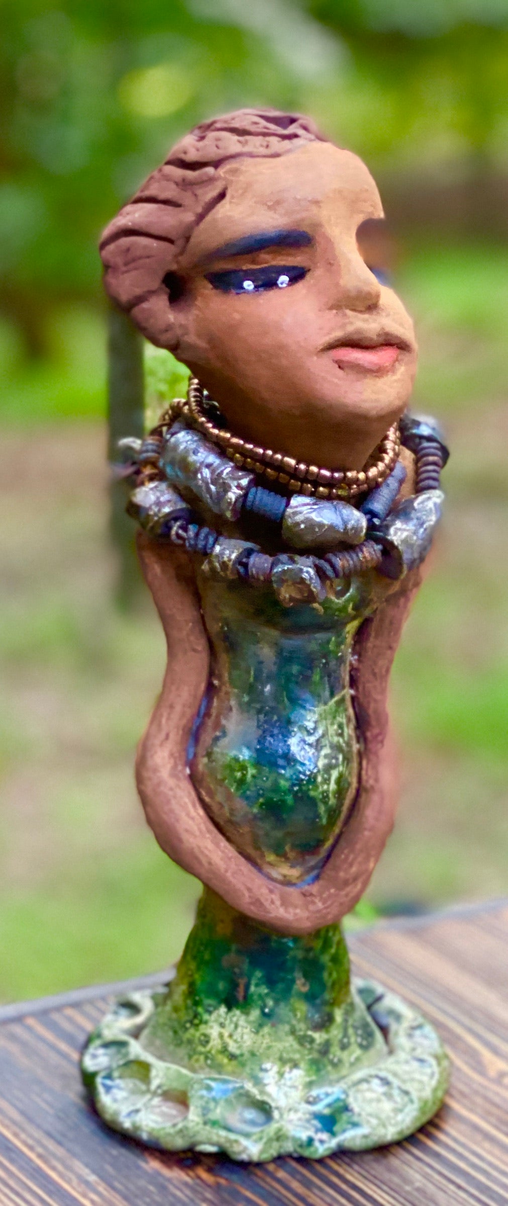 Greta! Greta stands 11" x 4" x 4" and weighs 2.5 lbs. She has a lovely honey brown complexion with  reddish brown lips. She has a braided hairstyle.  Gretaya has a colorful metallic green antique copper glazed dress. She wears two raku beaded necklaces. With long eyes slightly opened, Greta has hopes of finding a new home.