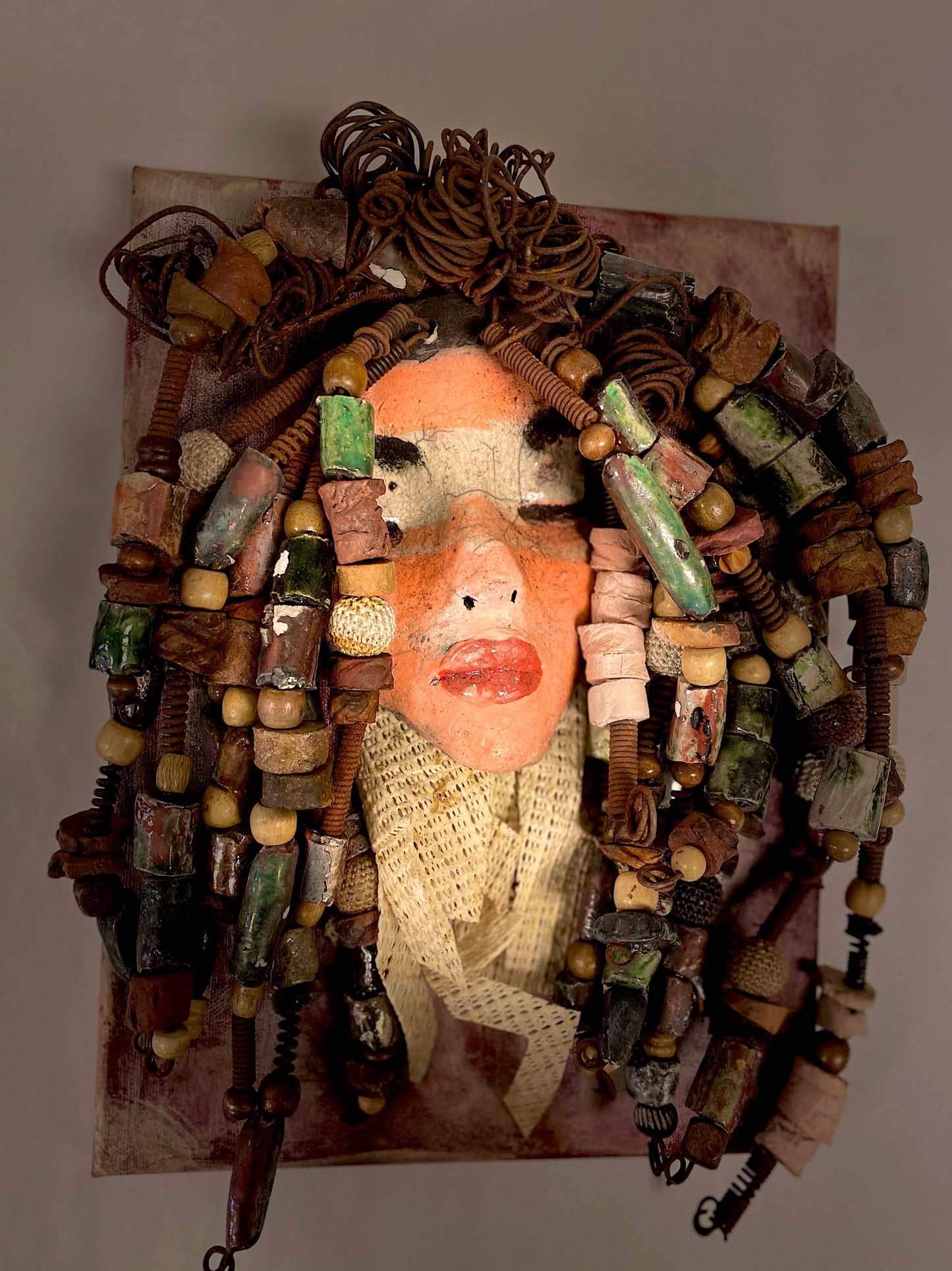 Daya is an exotic mask mounted on a painted 9"x 12" canvas. She weighs 1.9lbs. Daya's face is formed out of clay and raku fired. She has over 30 feet of hand coiled 16 gauge wire, over 50 hand formed raku beads, multiple wood beads, natural river stones, with a textured beige cloth. Daya's face beams with light pink and white matte and ruby red lips. She is ready to be hung!