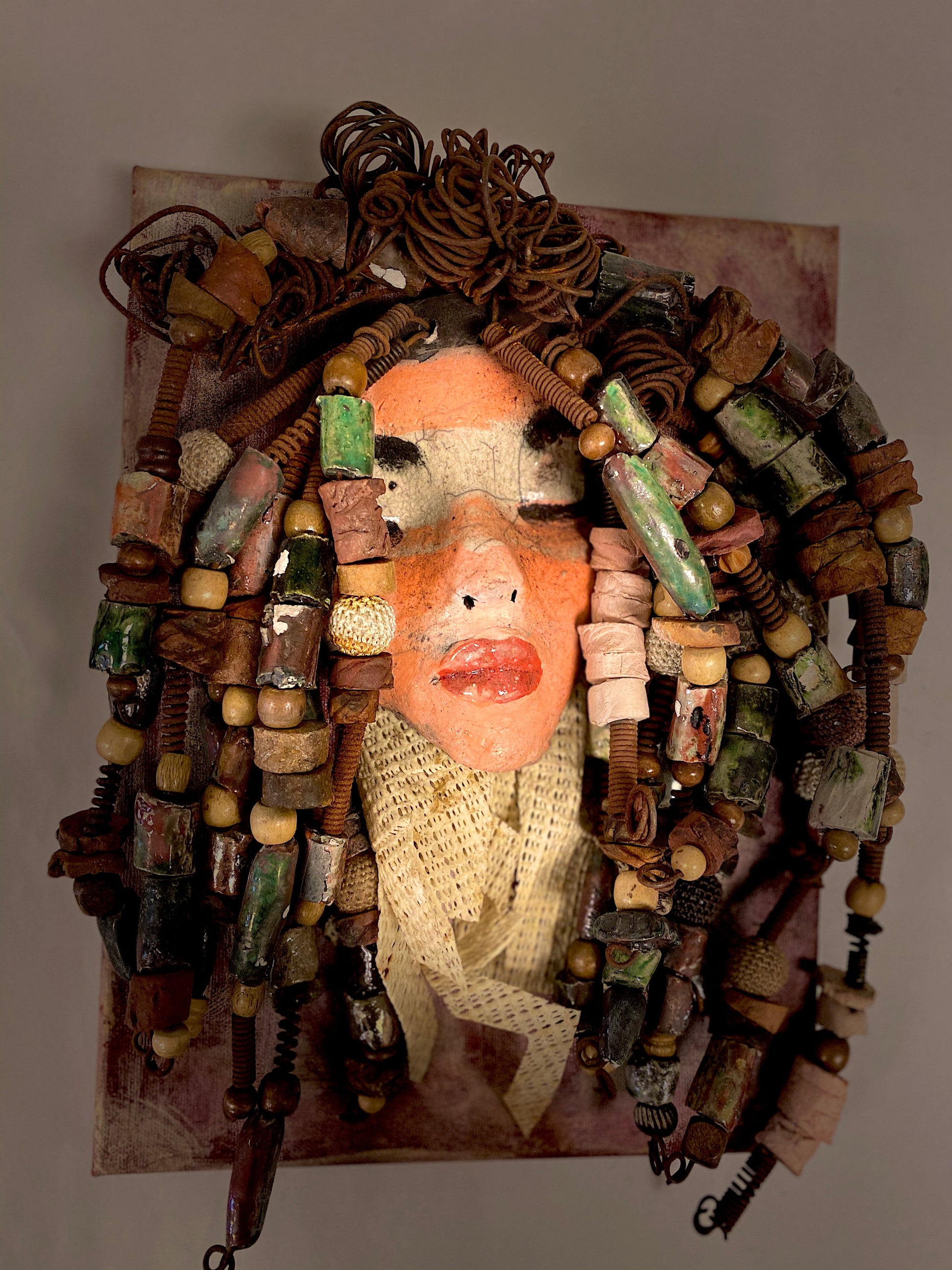 Daya is an exotic mask mounted on a painted 9"x 12" canvas.  She weighs 1.9lbs.  Daya's face is formed out of clay and raku fired.  She has over 30 feet of  hand coiled 16 gauge  wire, over 50 hand formed   raku   beads, multiple wood beads, natural river stones, with a textured  beige cloth.  Daya's face beams with light pink and white matte and ruby red lips.  She is ready to be hung!