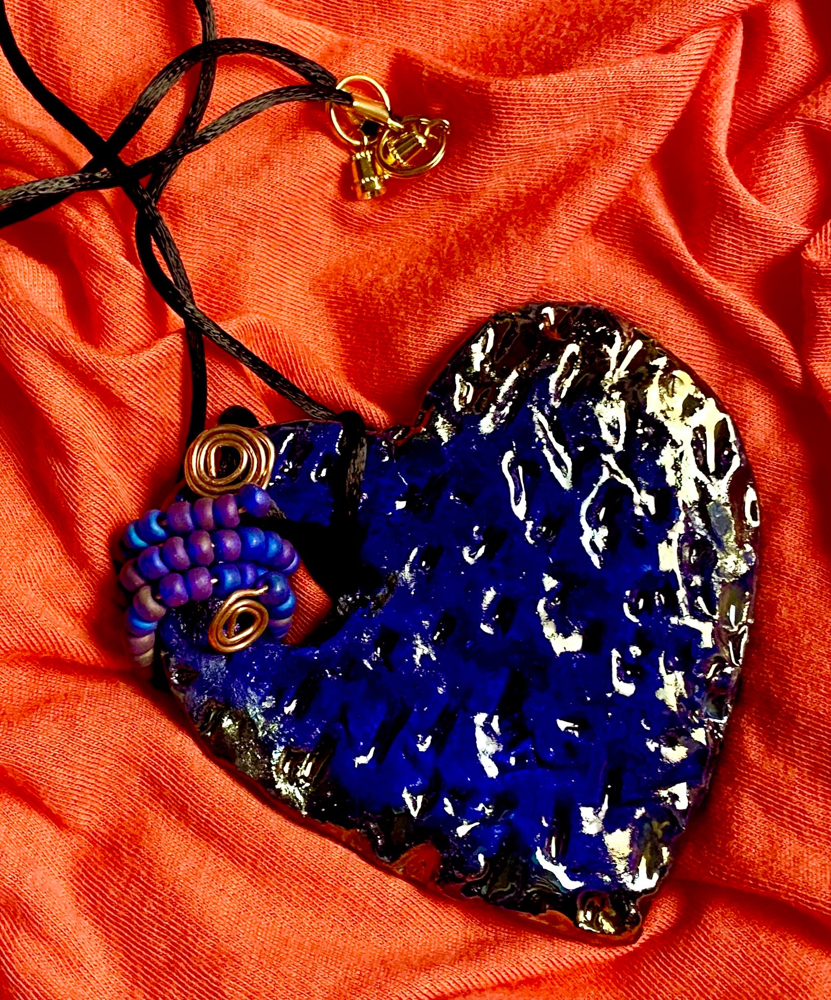 Have A Heart ! Each heart pendant is handmade with love! It is 3"x 3" and weighs approx. 3ozs. This pendant has a royal blue and gold metallic raku glazes that renders a unique translucent  patina. The heart  has a textured  pattern. It holds a spiral of off blue violet mini beads on a spiral copper wire. This pendant has a nice 12" black suede cord!