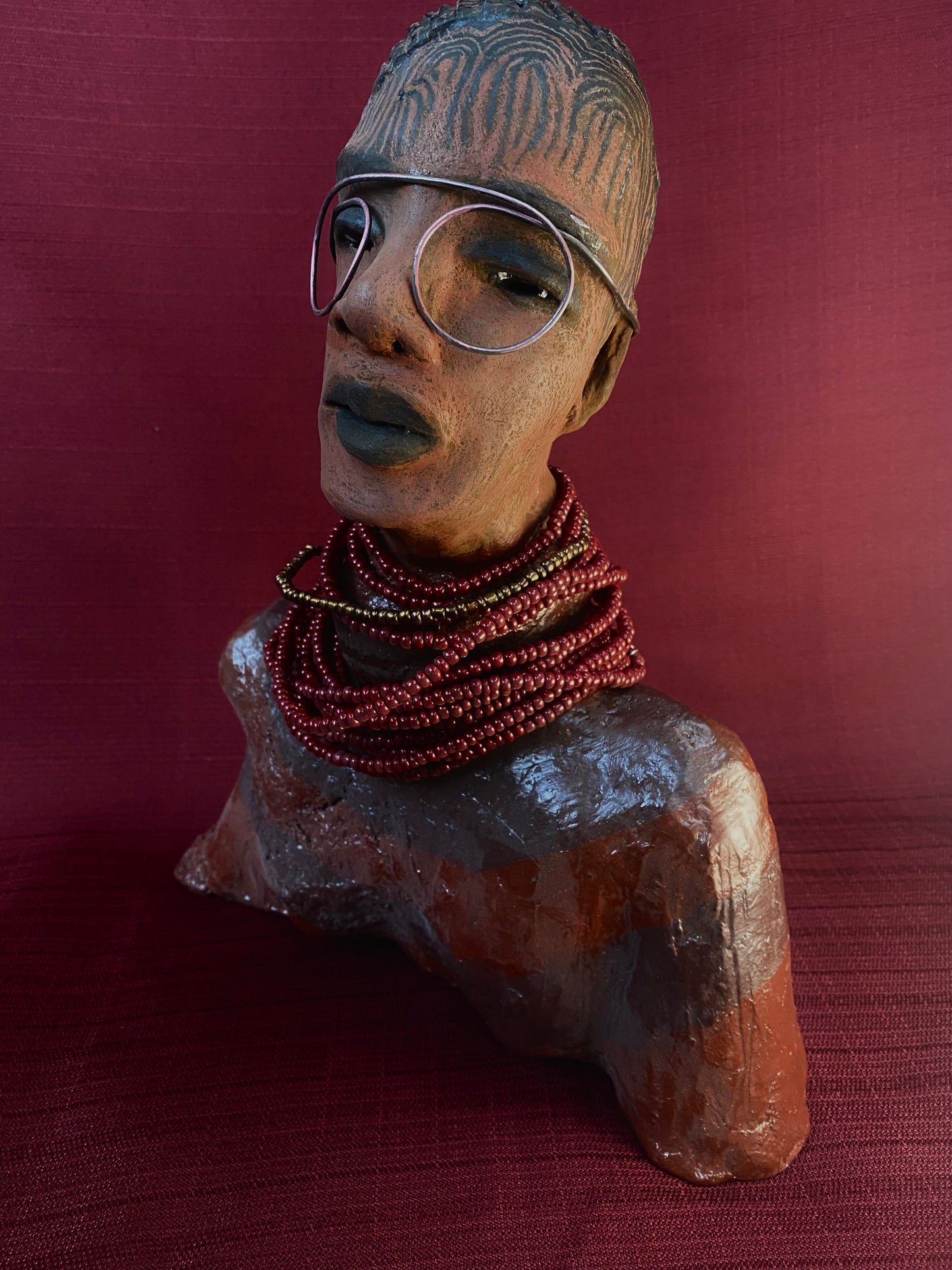 Meet Harriet! Harriet stands 13" x 11" x 5" and weighs 5 lbs. She wears a coral striped dress with a string of ruby red and gold beads. Harriet has a honey brown rust complexion and distinct eyes. Her eyes are slightly opened underneath her glasses... She has mixed black and copper streaks of straight corn rolls of clay hair. She has a straight look of concern as to what is about to take place.