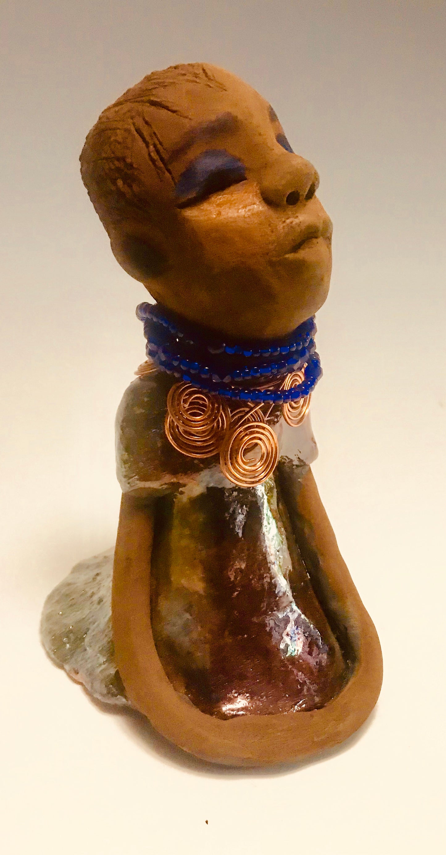Sade stands 7.5" x 5" x 5" and weighs 1.04 lbs. She has a lovely honey brown complexion with reddish brown lips. She has a short braided hairstyle. Sade has a colorful metallic antique copper glazed dress. She wears spiral copper wire necklaces on top of an aqua blue beaded collar. Sade long loving arms rest at her side. With accent blue eye shadow and eyes wide opened, Sade has hopes of finding a new home.