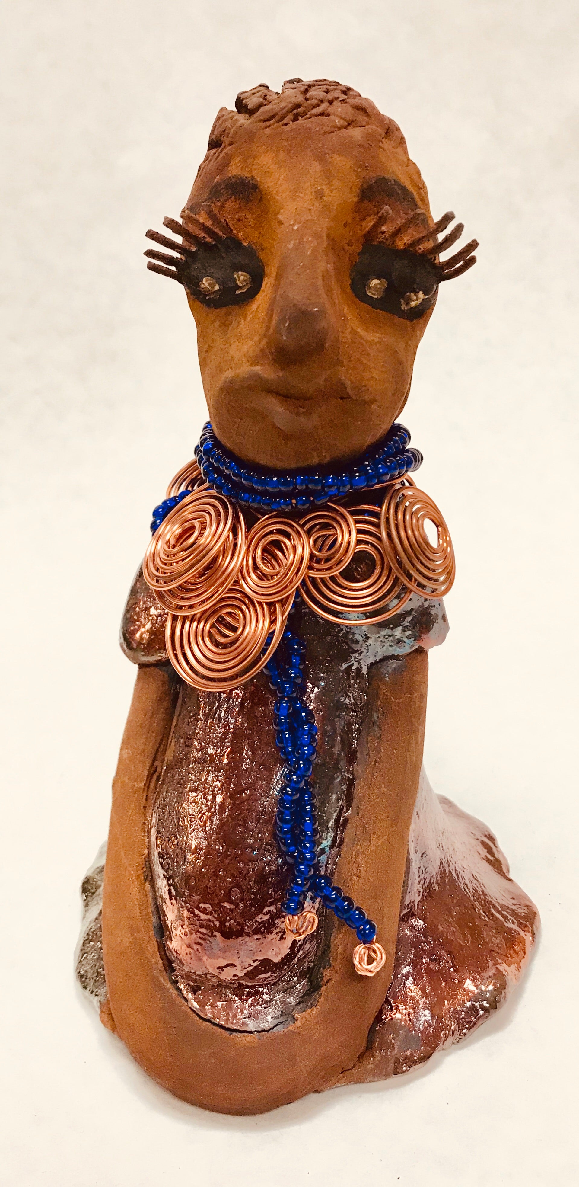 Tattayana stands 8" x 4.5" x 5" and weighs 1.02  lbs. She has a lovely honey brown complexion with reddish brTattayanaown lips. She has a short braided hairstyle.  Tattayana has a colorful metallic antique copper glazed dress. She wears spiral copper wire necklaces on top of an aqua blue beaded collar.  Tattayana has her long loving arms rest at her side. With eyes wide opened,Tattayana  has hopes of finding a new home.